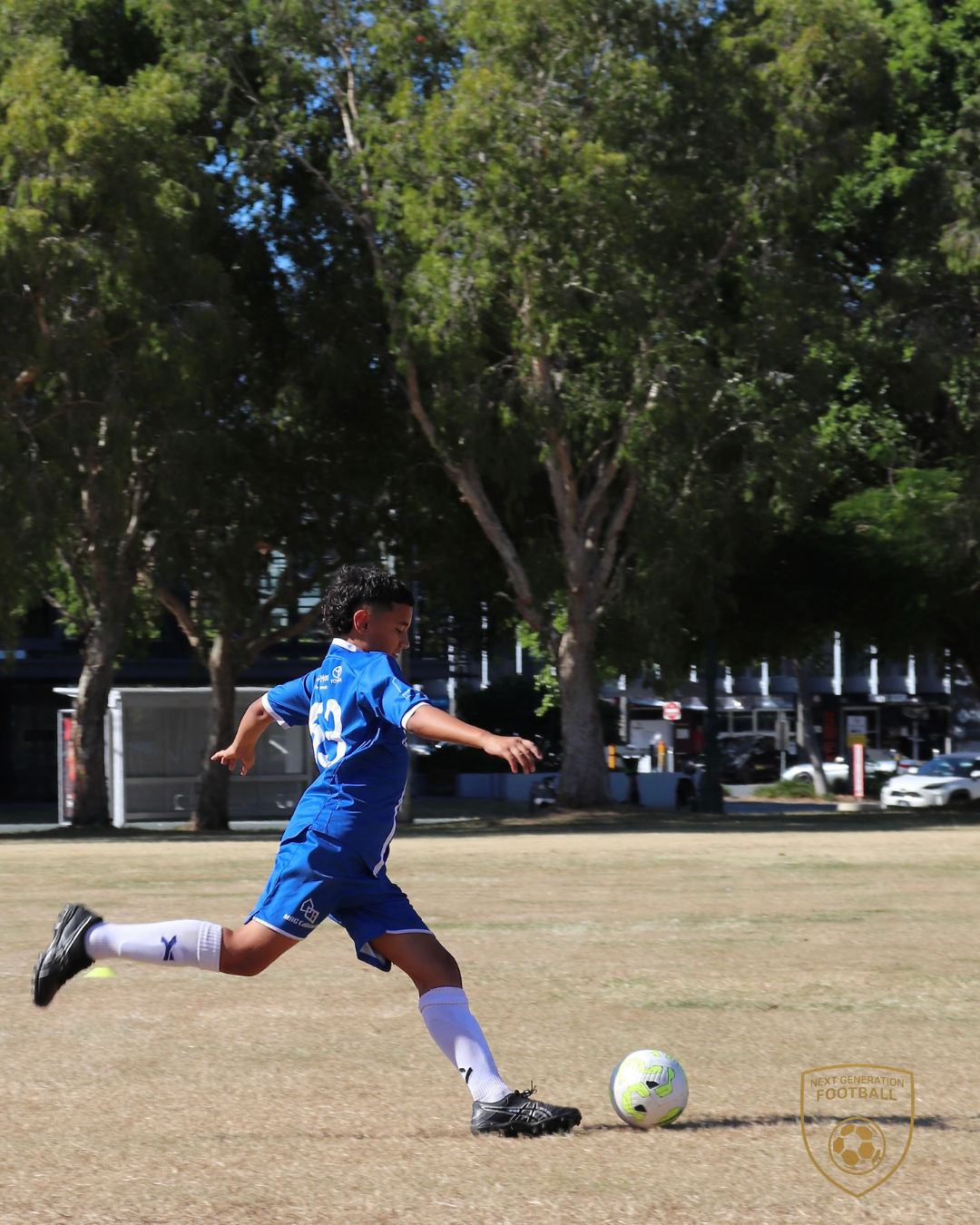 Young boy in a blue soccer uniform kicking a soccer ball on a grassy field with trees and parked cars in the background.