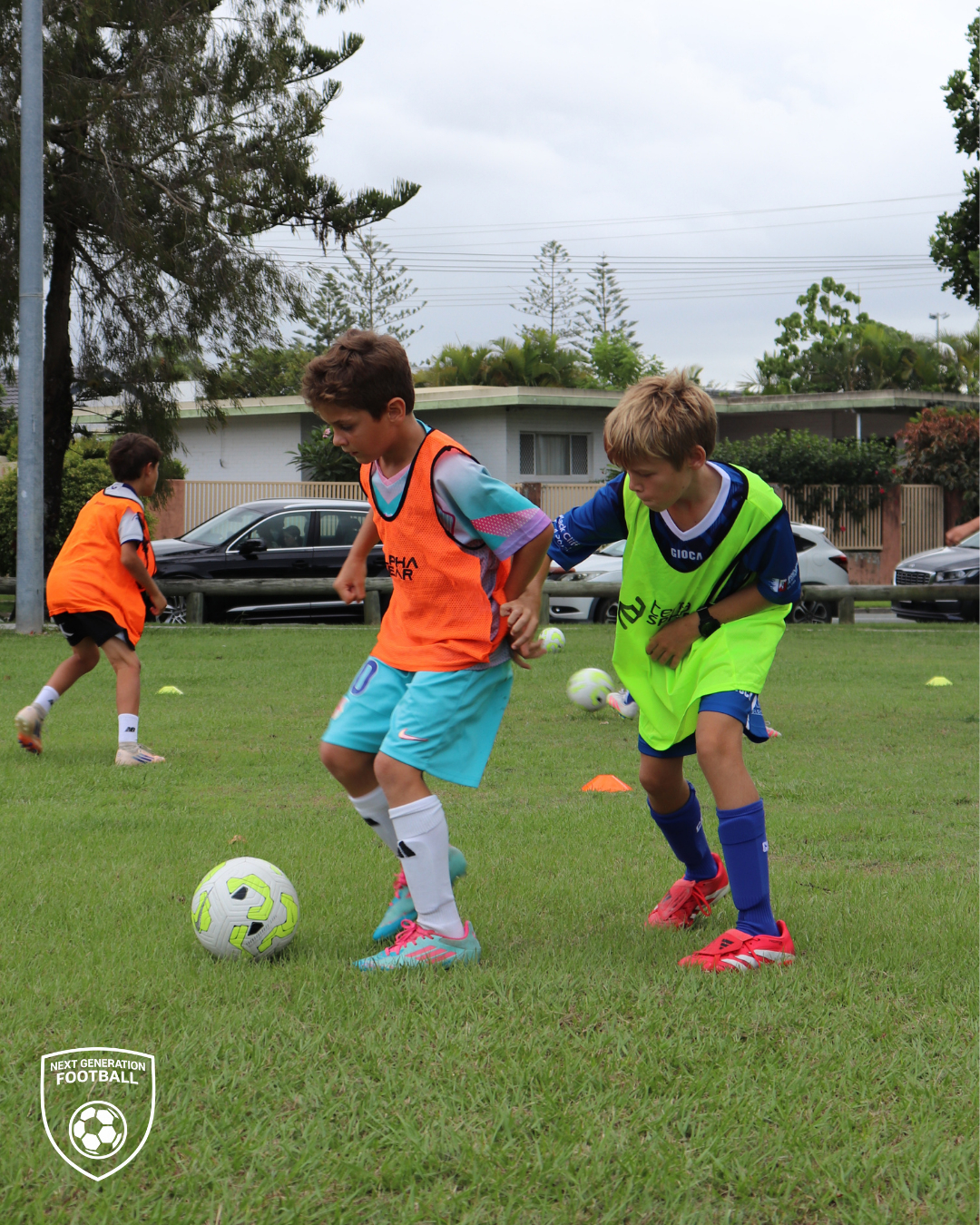 Young boys playing soccer on a grassy field during daytime, wearing colored pinnies, with trees, houses, and parked cars in the background.