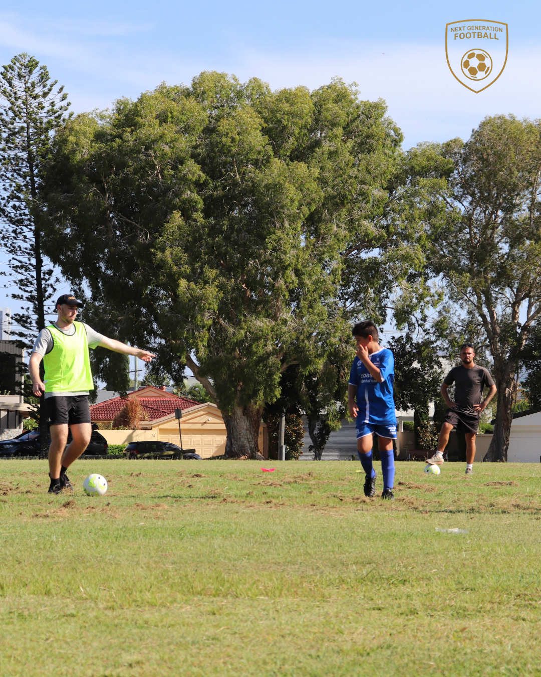 A coach and two young soccer players practicing drills on a grassy field with trees and houses in the background.
