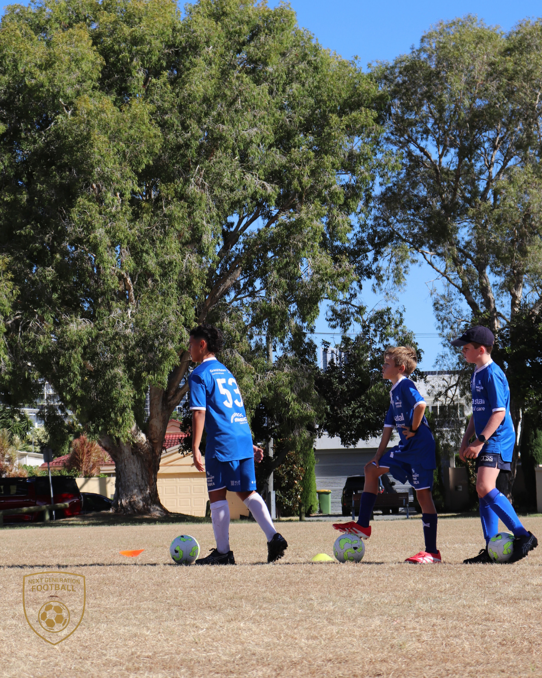Three young boys in blue soccer uniforms practicing dribbling on a soccer field with trees in the background.