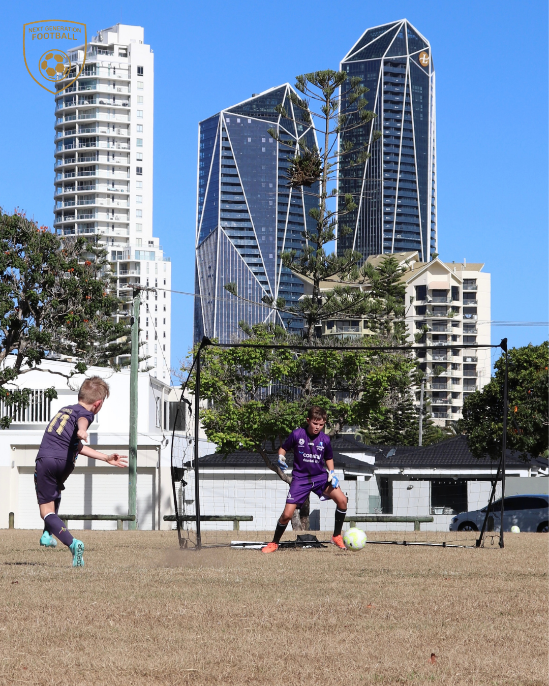 Two young boys playing soccer in a backyard with a portable goal, with tall modern buildings, trees, and a bright blue sky in the background.