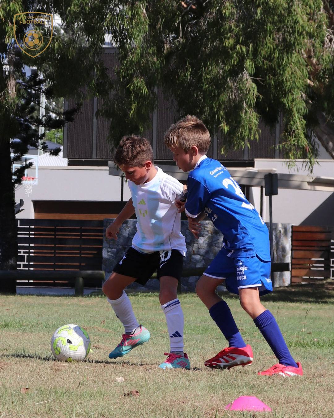 Two young boys play soccer outdoors on grass, with one controlling the ball and the other defending. The boy in white has light-colored cleats, white socks, and black shorts, while the boy in blue has bright red cleats, blue socks, and blue shorts. A