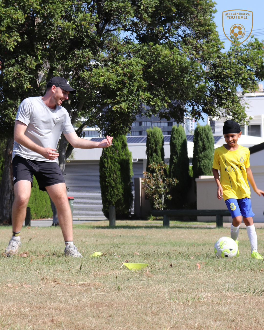 A man and a young boy playing soccer on a grassy field in a park, with trees and buildings in the background.