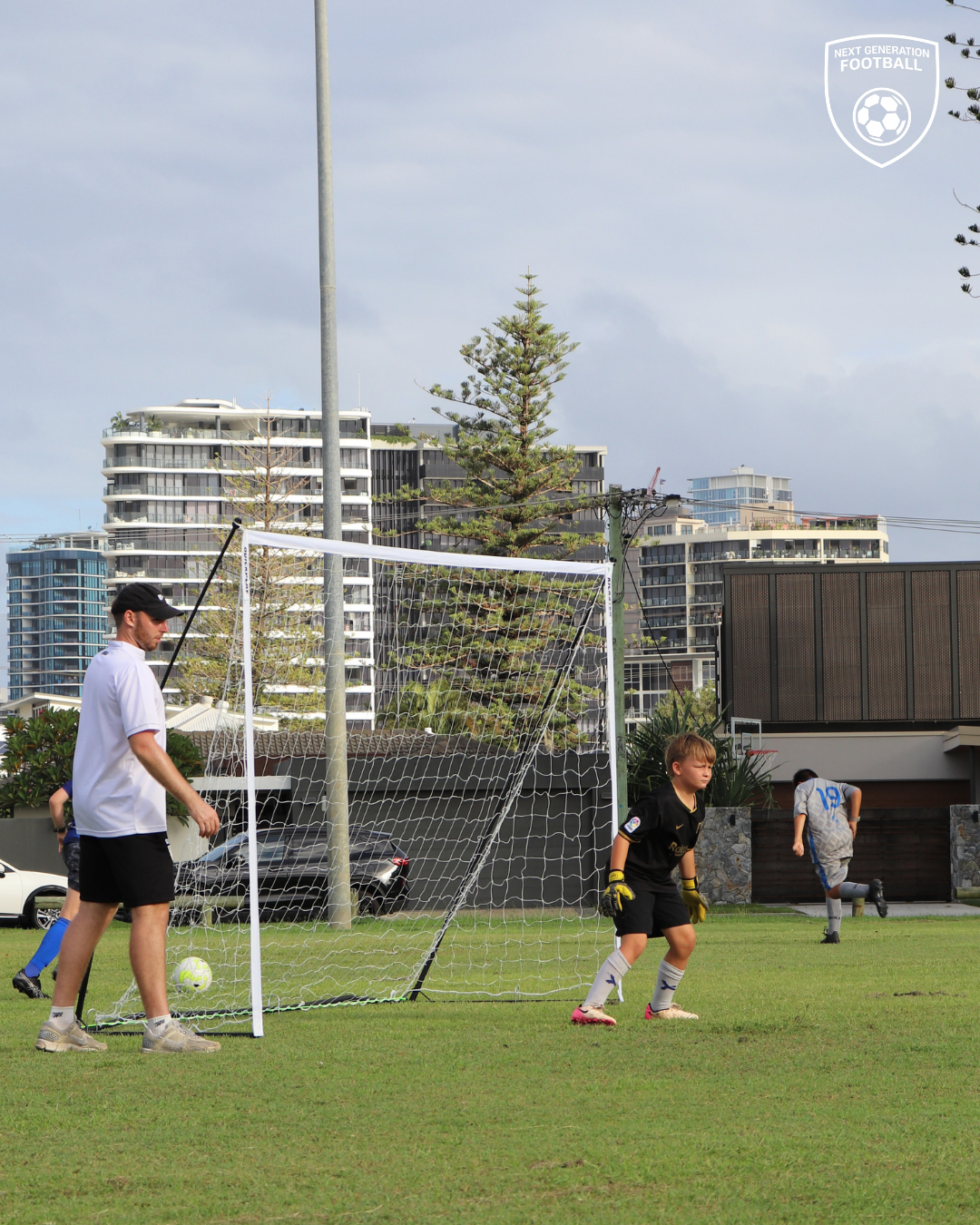 Kids playing soccer on a grassy field, one kid acting as goalkeeper, others nearby, multi-story buildings in the background.