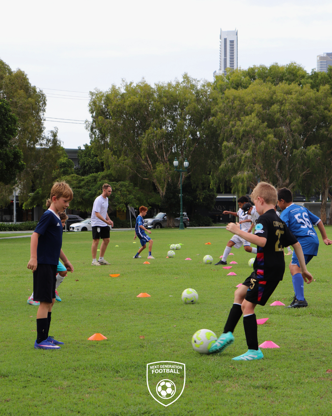 Children participating in a youth soccer practice on a green field with training cones and multiple soccer balls, overseen by an adult coach, with trees and tall buildings in the background.
