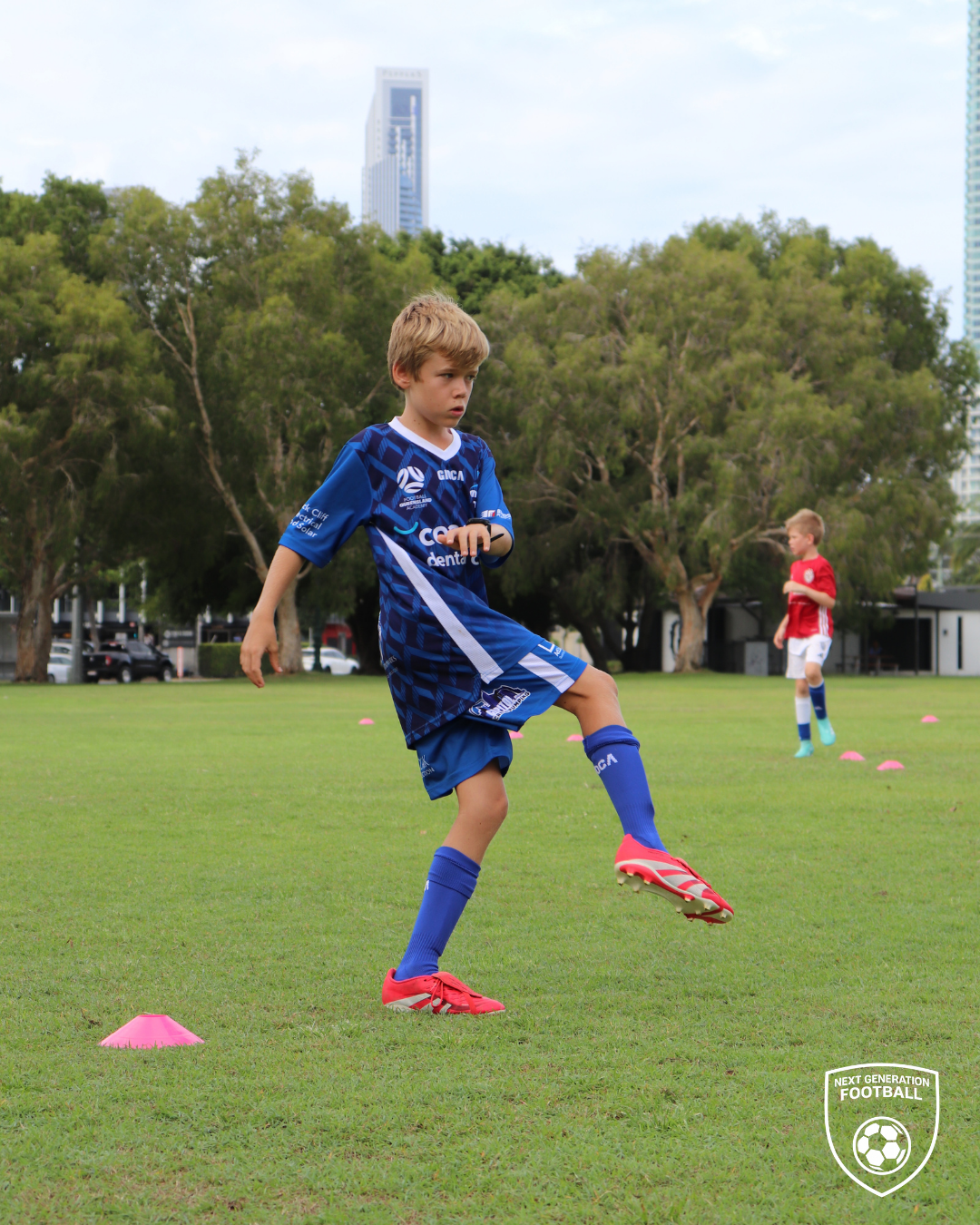 Young boys practicing soccer on a grassy field with pink training cones, one in a blue jersey kicking a ball, and the other in a red jersey standing in the background, with trees and tall buildings in the background.