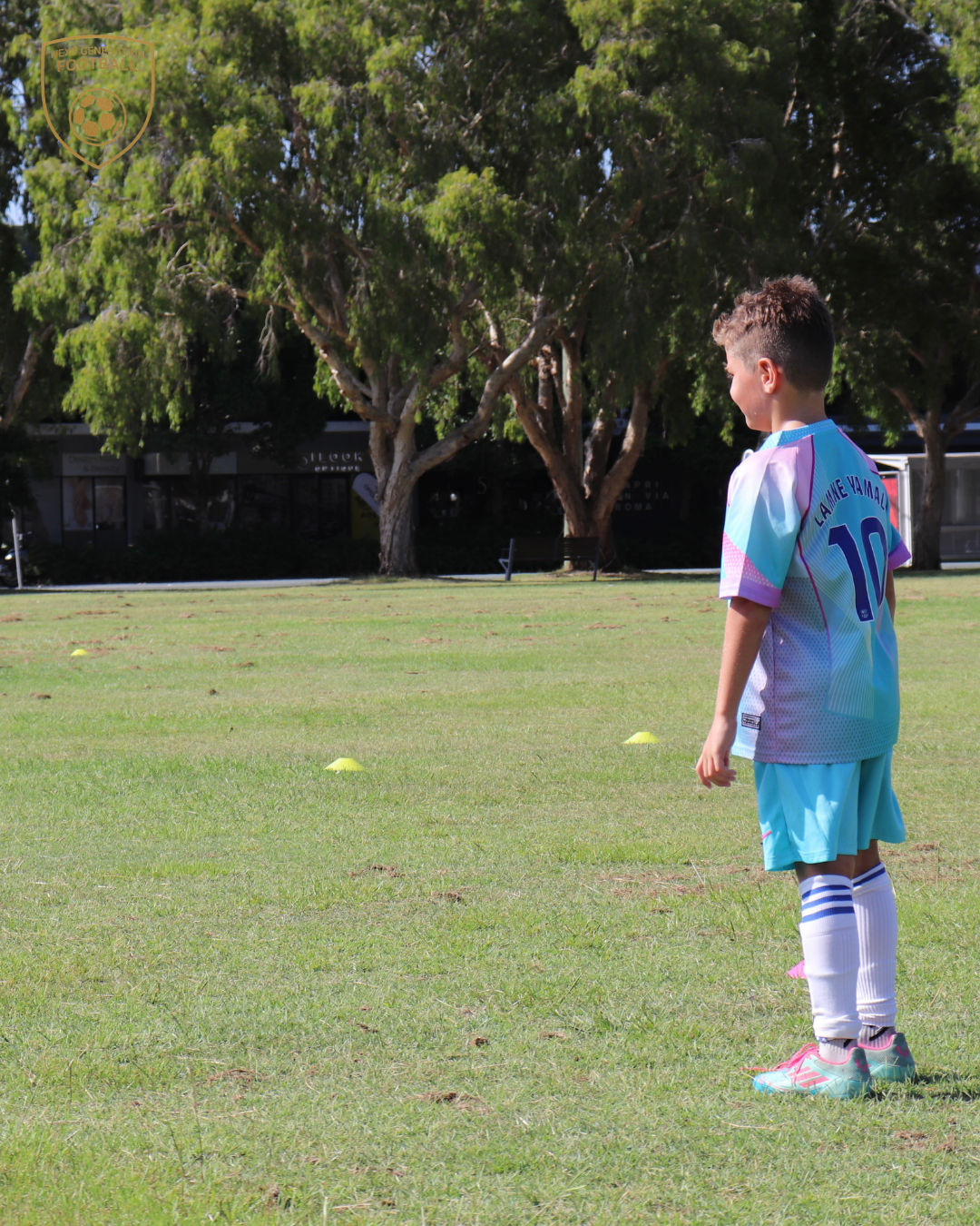 A young boy in a soccer uniform standing on a grassy field with small yellow cones scattered around, with trees and buildings in the background.