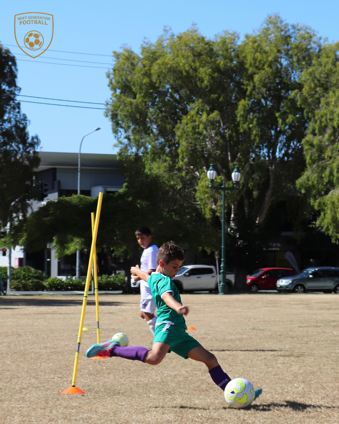 Young boys practicing soccer on a field with cones and poles, with trees and parked cars in the background.