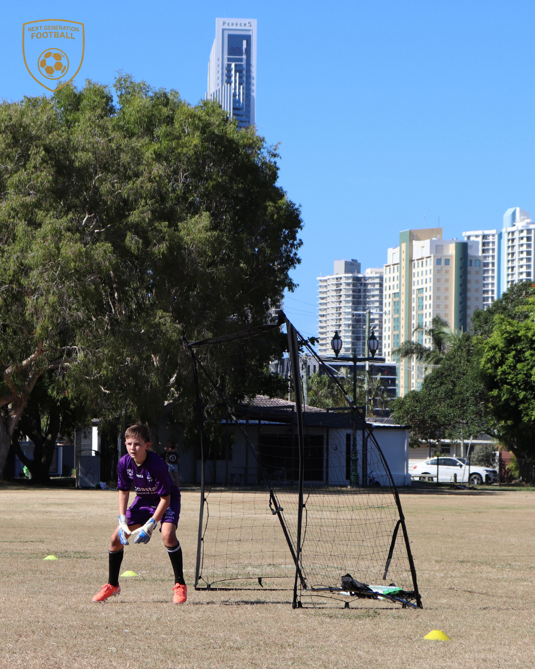 A young boy playing soccer as a goalkeeper on a grassy field with city buildings in the background, sunny sky, and trees around.