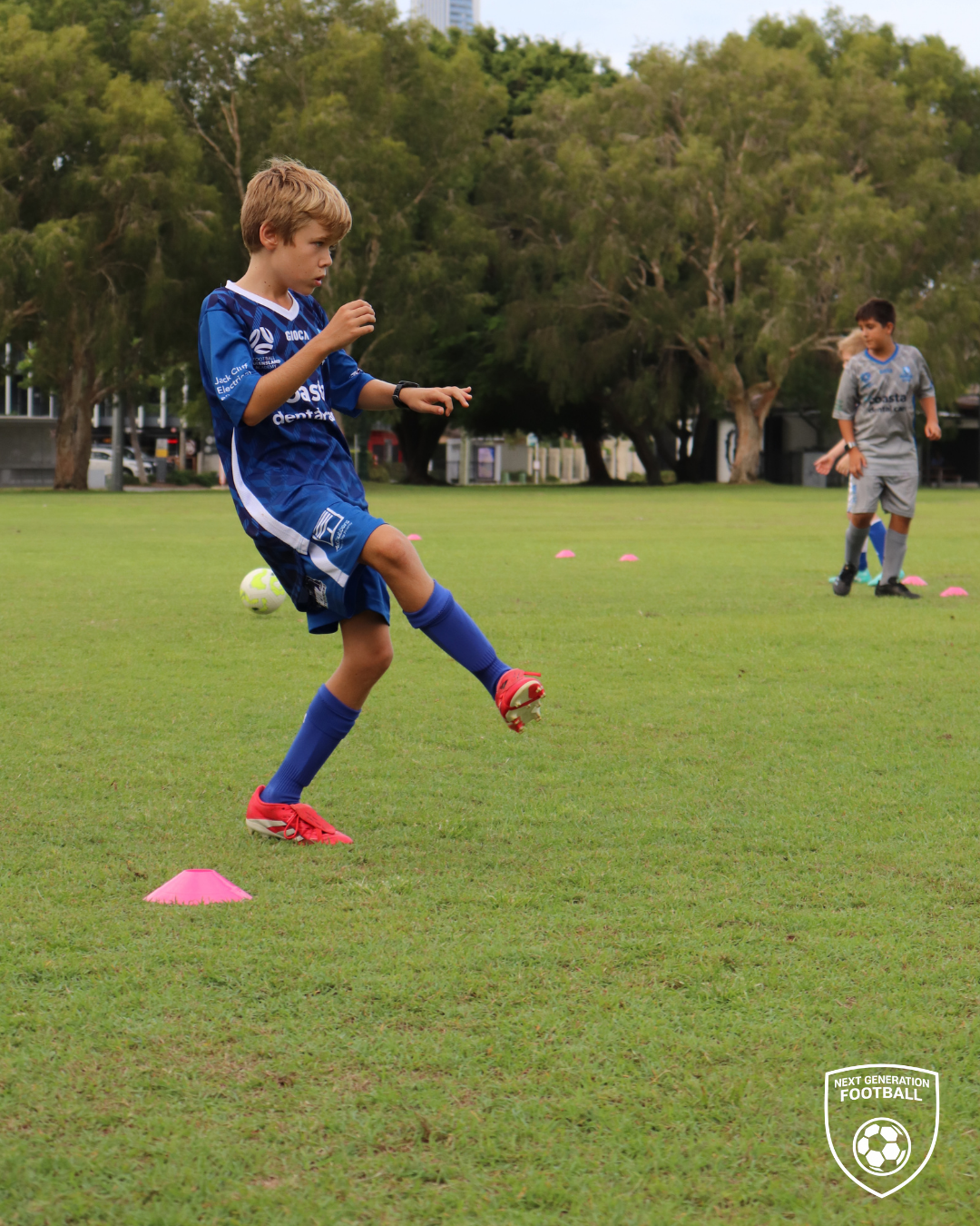 Child in blue soccer uniform practicing dribbling around pink cones on a grassy field, with trees and another child in gray uniform in the background.