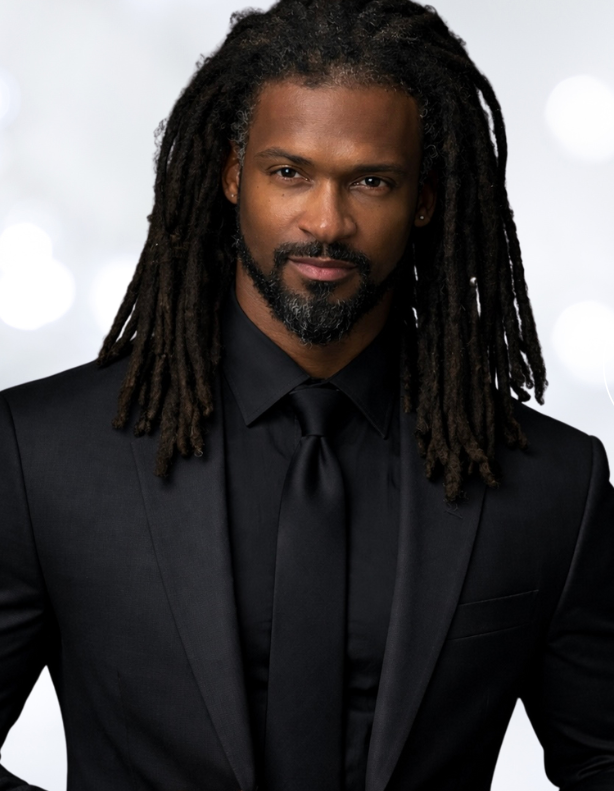 A man with long dreadlocks, a beard, and mustache wearing a black suit, black shirt, and black tie, standing against a blurred background with circular light reflections.