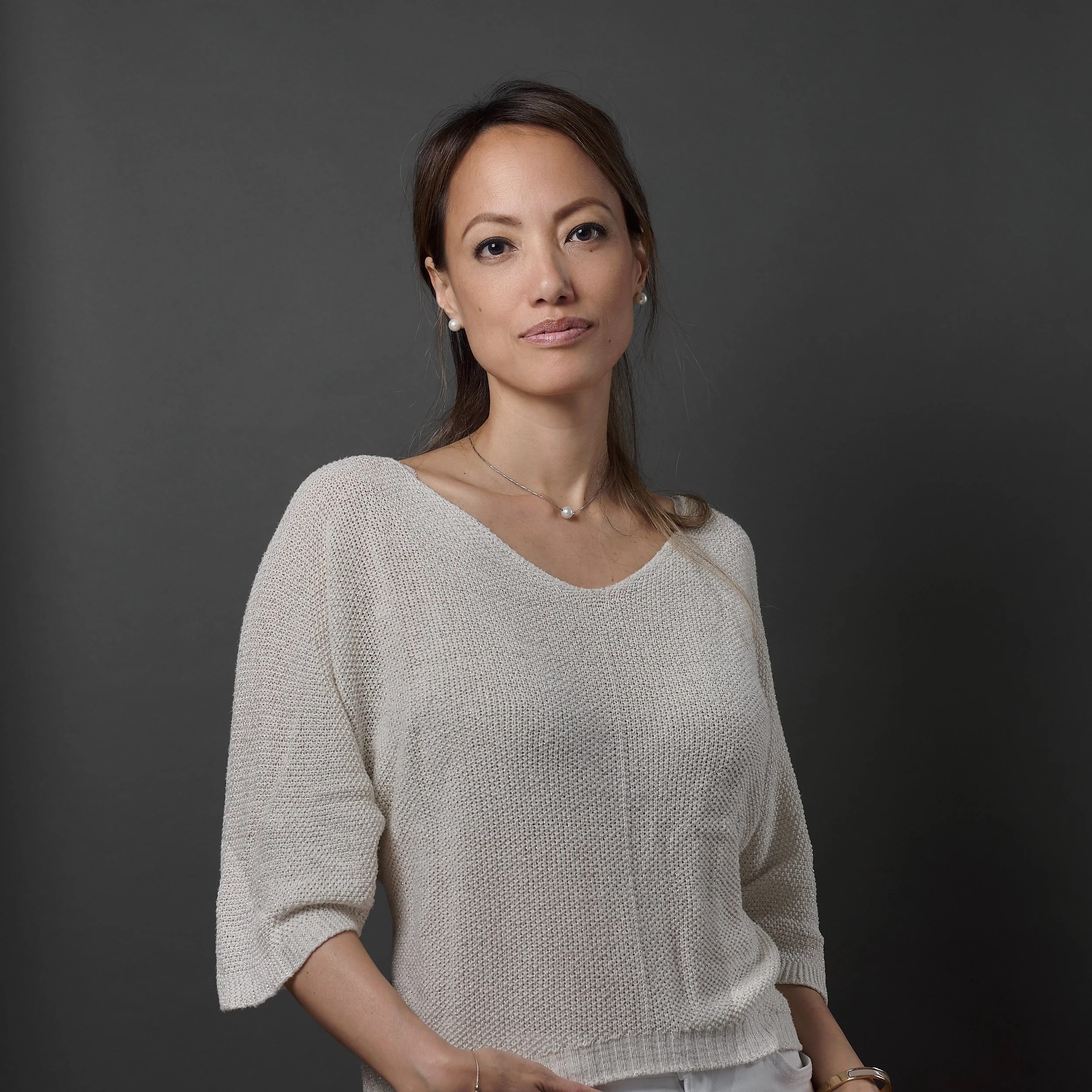 A woman with light skin and brown hair styled in a loose updo, wearing pearl earrings, a pearl necklace, a beige mesh top with three-quarter sleeves, posing against a plain gray background.