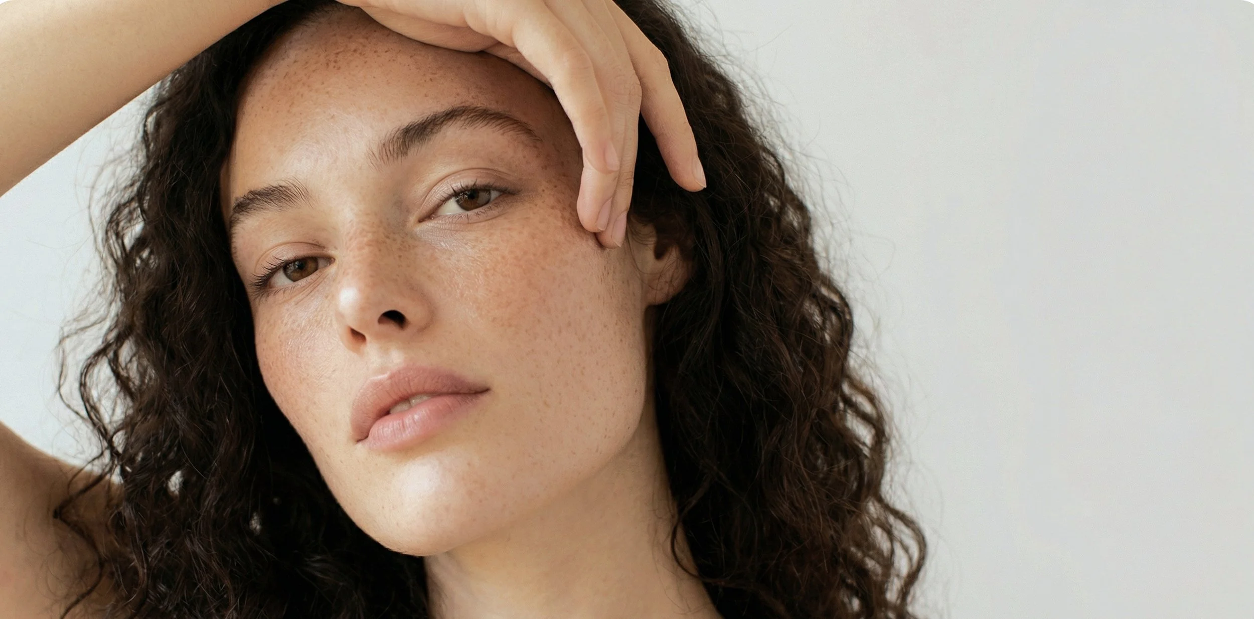 A woman with curly brown hair and freckles resting her hand on her forehead, looking at the camera with a calm expression.