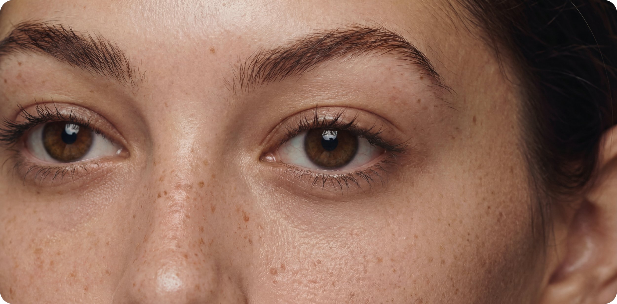 Close-up of a person's face showing brown eyes with makeup, freckles, and dark eyebrows.