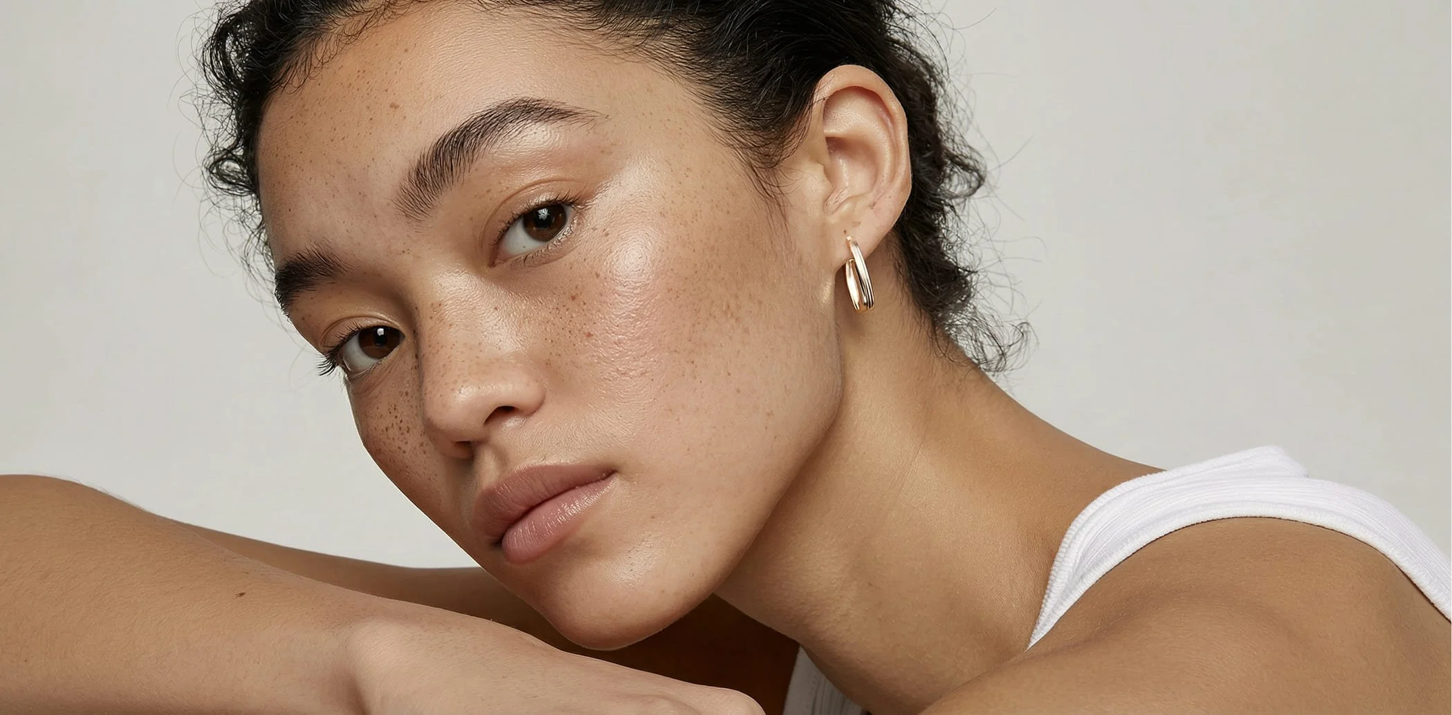 Close-up of a young woman with curly dark hair, brown eyes, and light brown skin with freckles, wearing a white tank top and a gold hoop earring, resting her arm on her shoulder, against a plain light background.