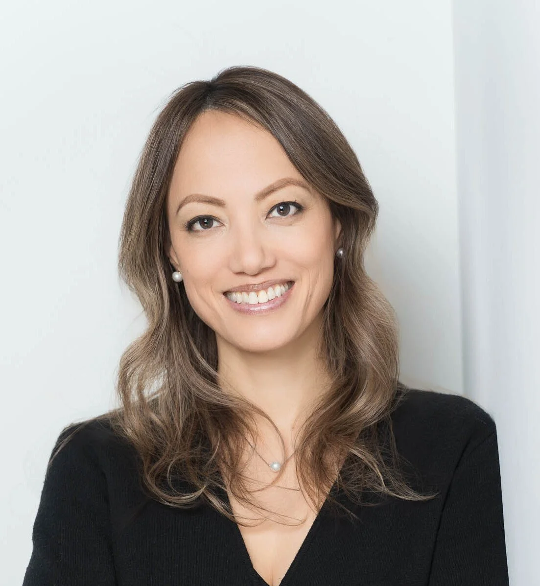 A woman with shoulder-length brown hair, smiling, wearing pearl earrings and a black top, standing against a white background.
