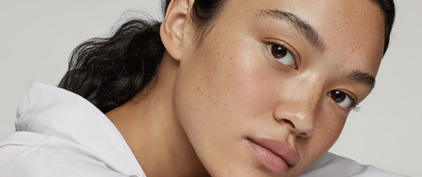 Close-up of a young woman with clear, freckled skin and dark hair, wearing a white shirt against a neutral background.