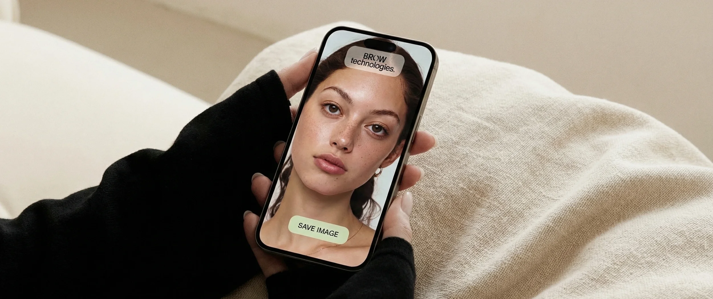 A person holding a smartphone displaying a photo of a young woman with freckles, brown hair, and hoop earrings, with the text 'BROW technologies.' and a button labeled 'SAVE IMAGE' on the screen.