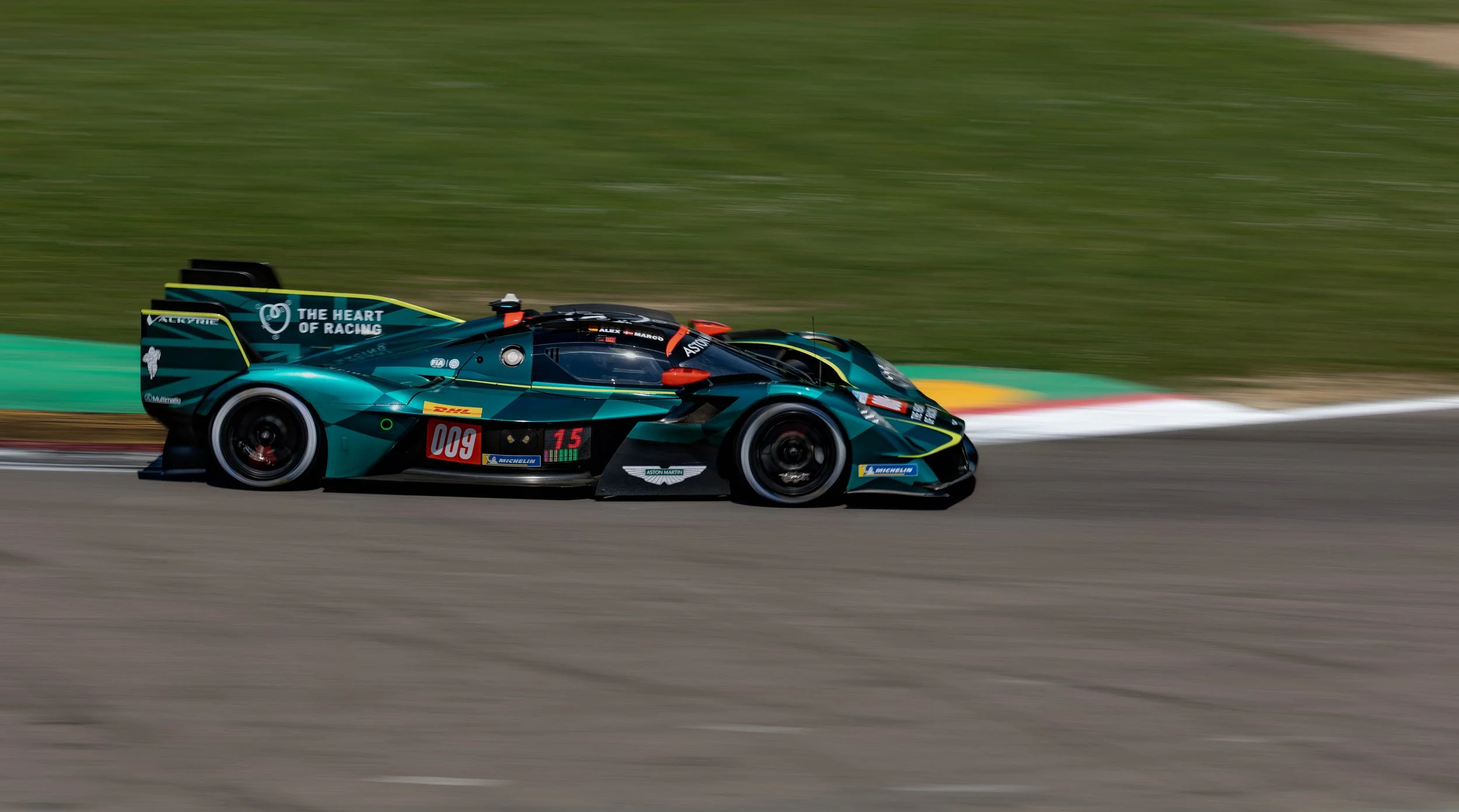 A race car with teal and black colors moving at high speed on a racetrack with a blurred green background.