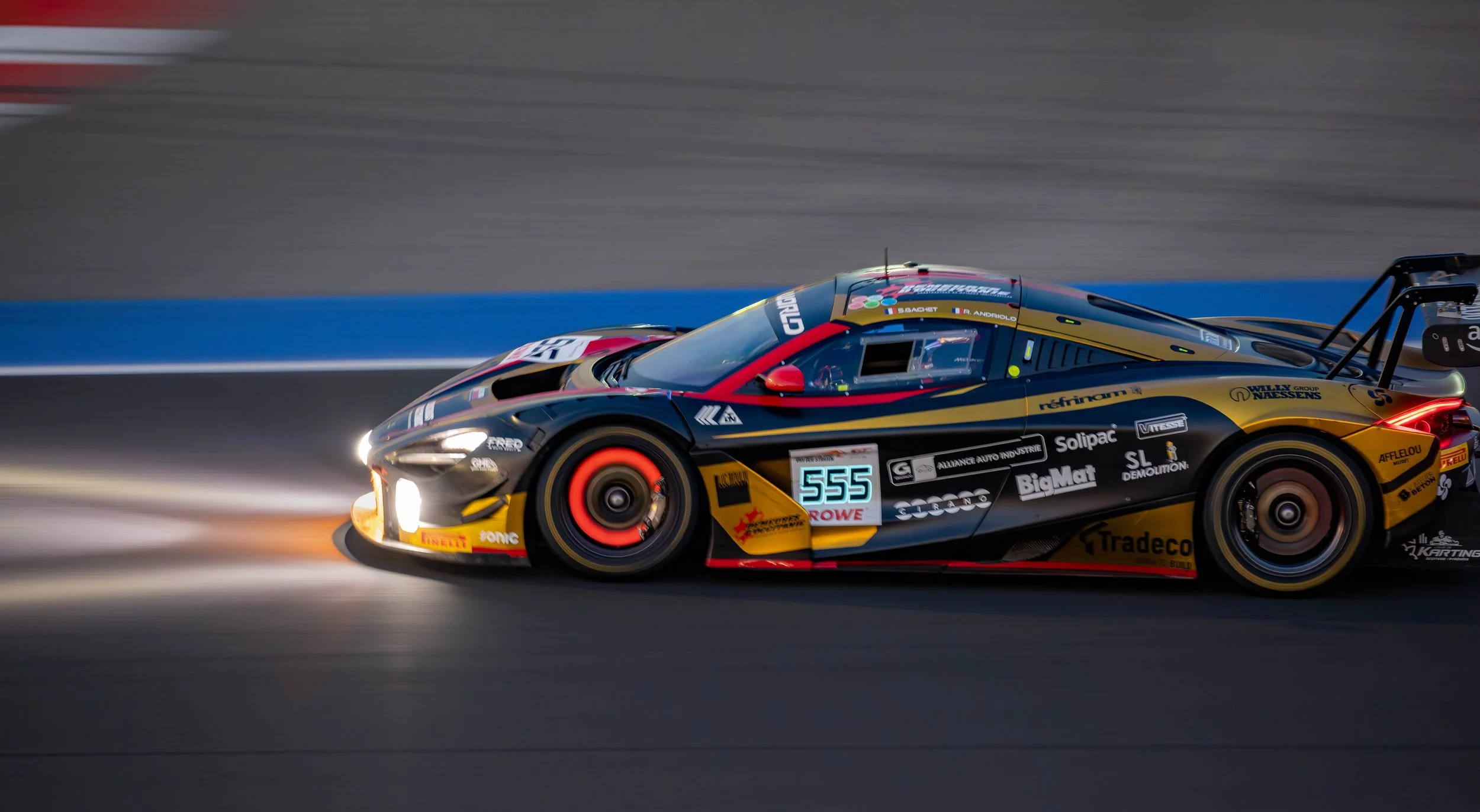 Race car moving quickly on track at night, illuminated headlights and colorful sponsor decals, blurred background.
