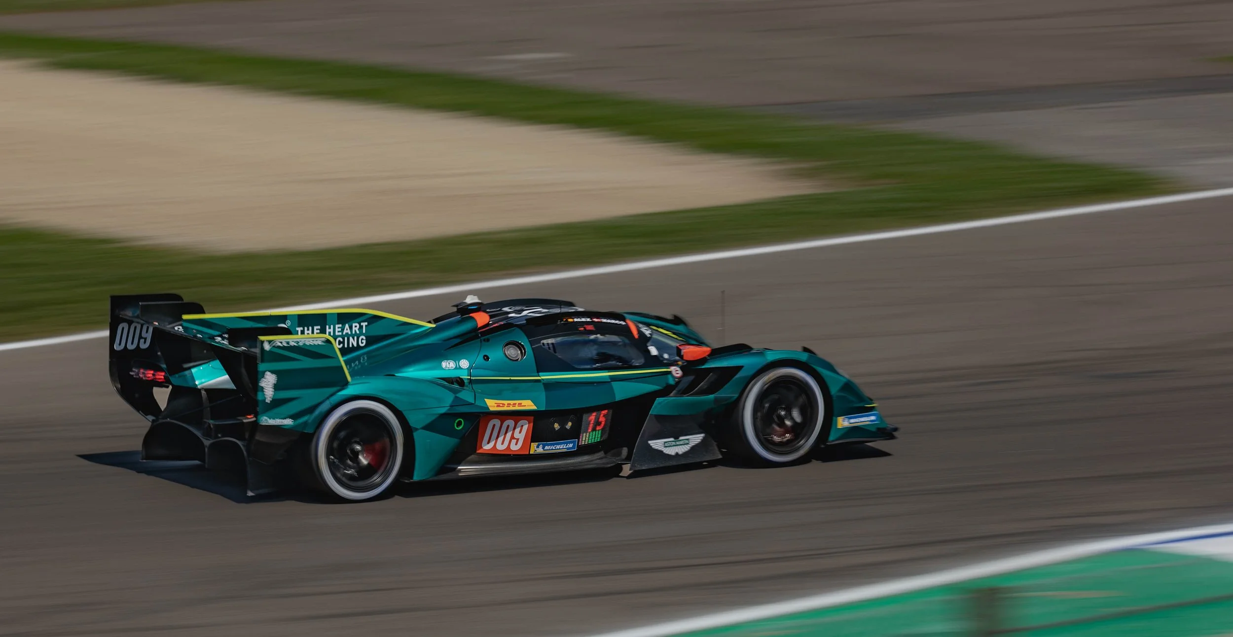 A teal and black race car on a race track during daylight, with sponsors and the number 009 on its side.