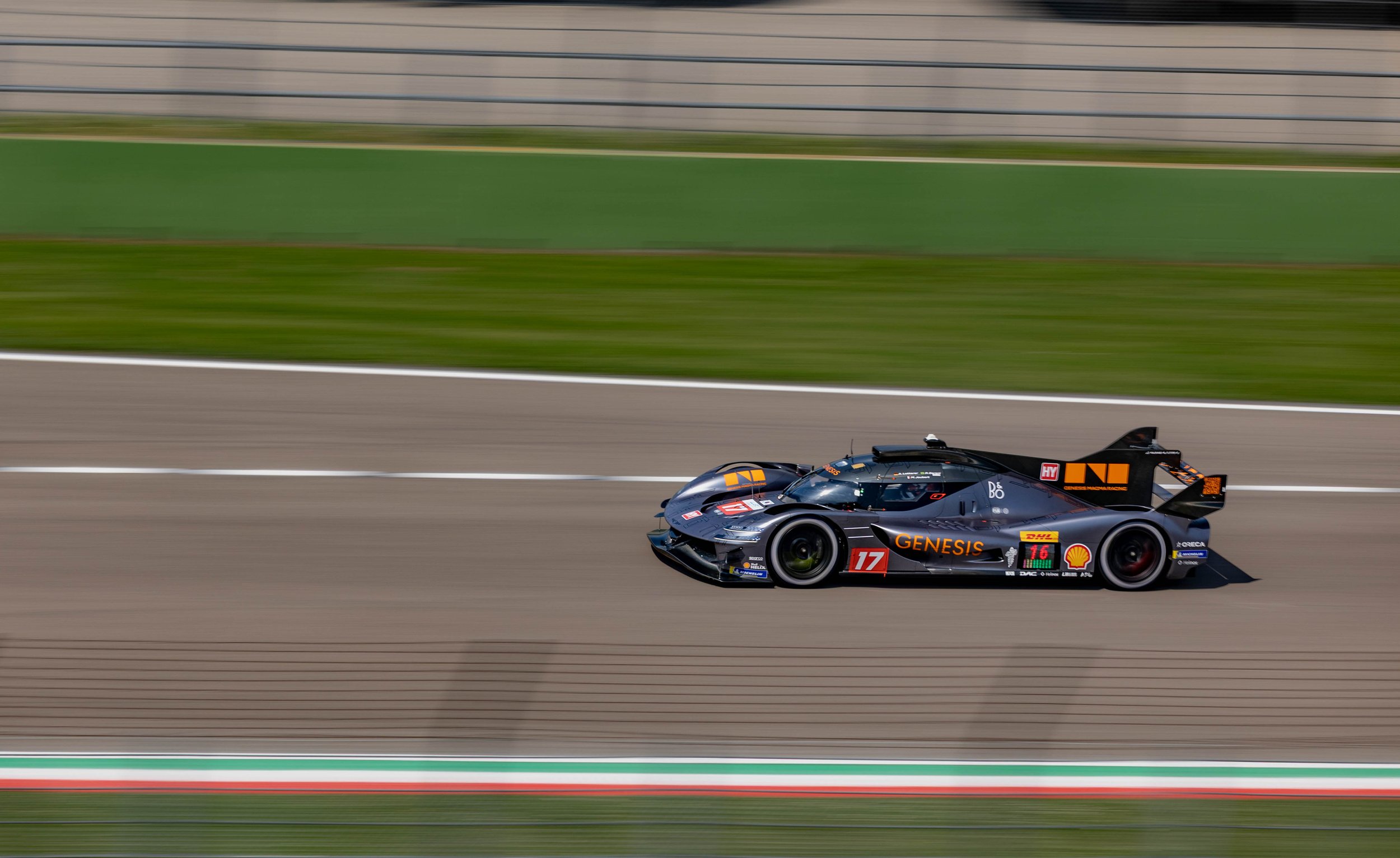 A black race car with sponsor logos speeding on a racetrack, with a motion blur background and green grass.