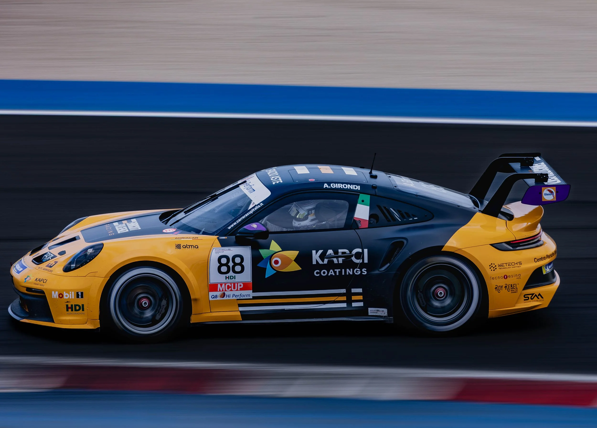 A yellow race car speeding on a racetrack, with various sponsor logos and the name A. Girondi on the windshield, in motion blur background.