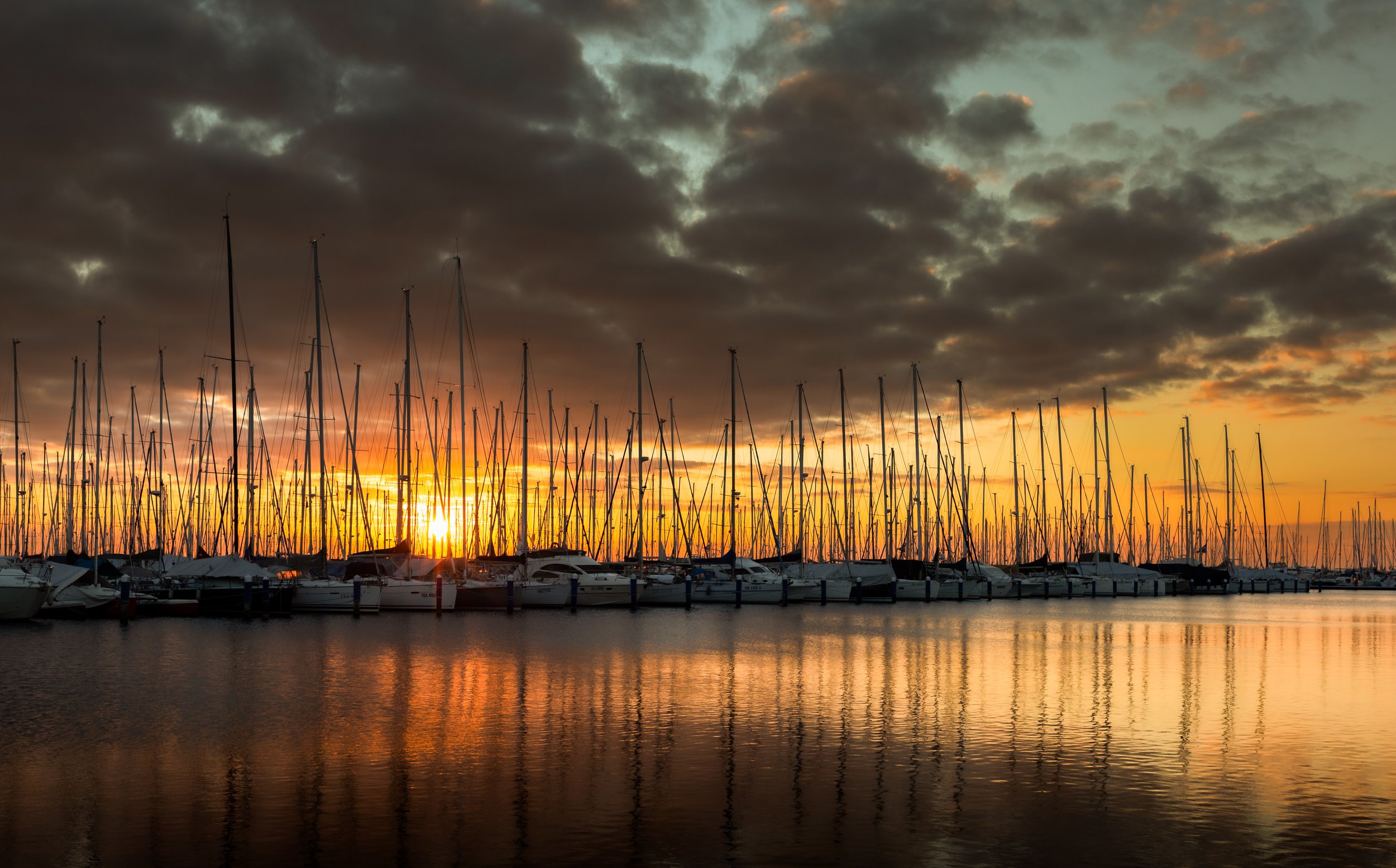 A marina with sailboats docked in calm water during sunset, with dark clouds overhead and the sun near the horizon casting an orange glow.