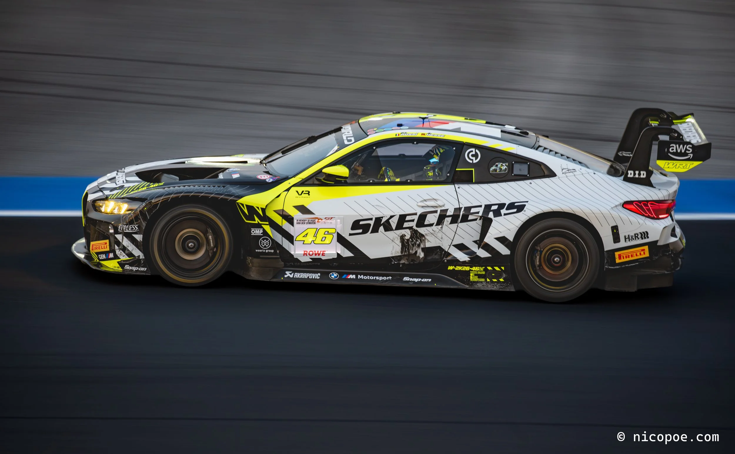A race car with black, white, and yellow livery speeds on a race track, with a blurred background indicating motion.