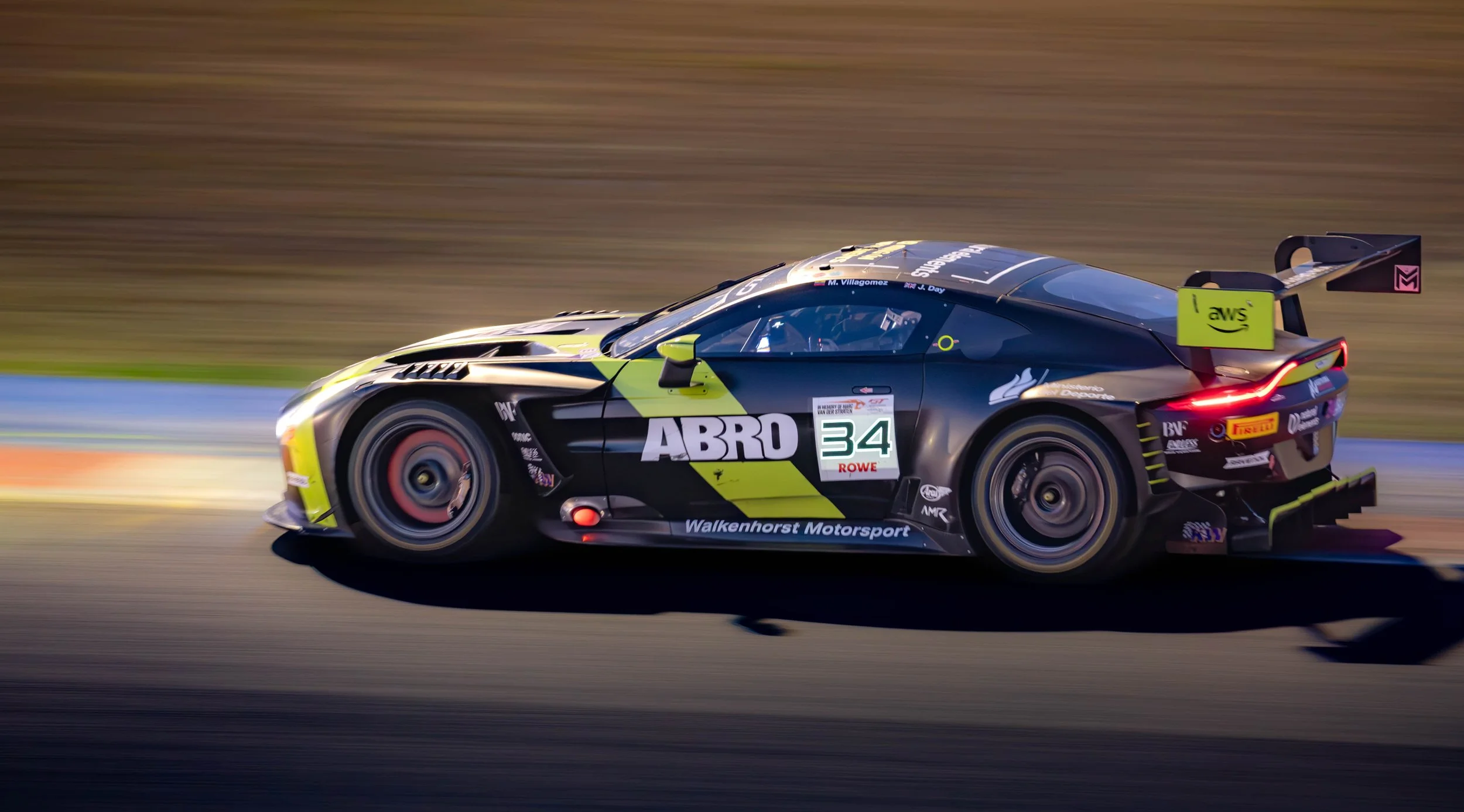 A black and yellow race car with the number 34 on the side, speeding on a race track. The car has sponsor logos, including ABRO, AWS, and Pirelli, and is in motion with blurred background and wheels.