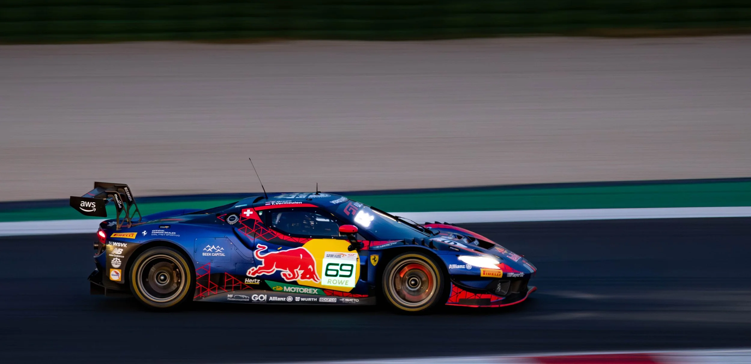 A racing car on a race track with a blurred background, featuring sponsorship logos, red and blue color scheme, and a large rear wing.