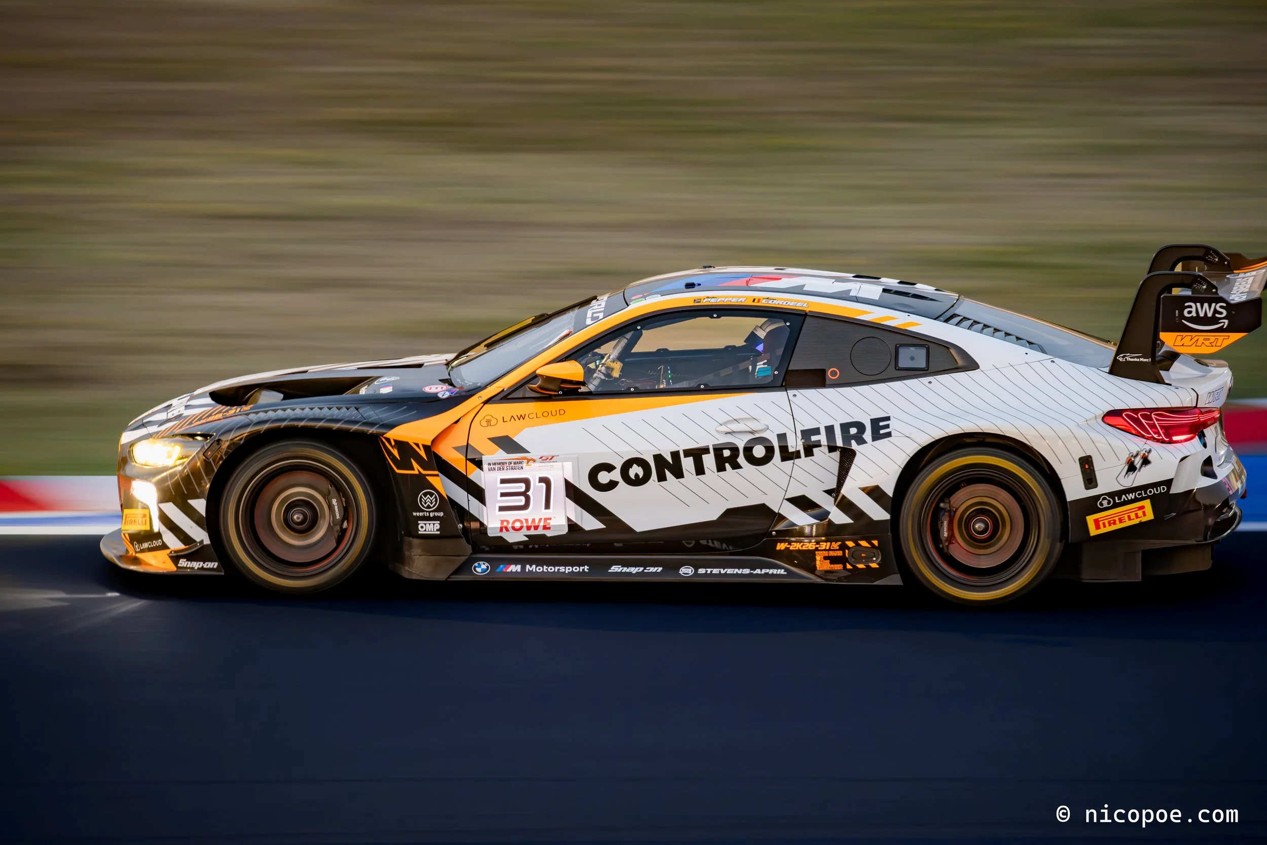 A race car speeding on a track with a blurred background. The car is black and white with orange accents and has multiple sponsor logos.