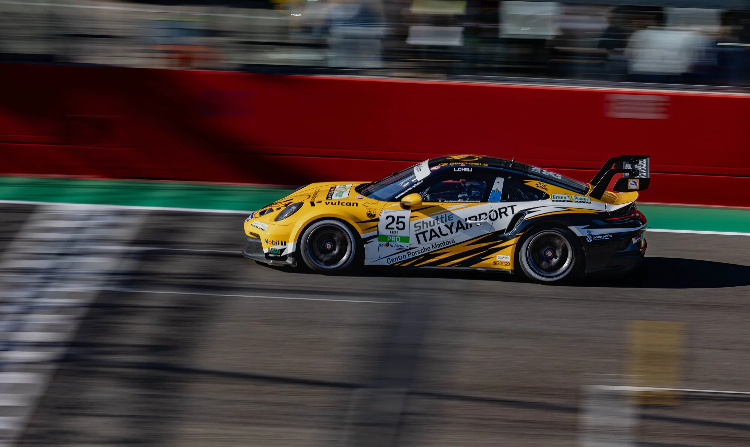 A yellow and black race car speeding down a racetrack with motion blur, with the number 25 on its side and various sponsor logos. The background shows a red barrier and blurred spectators or structures.