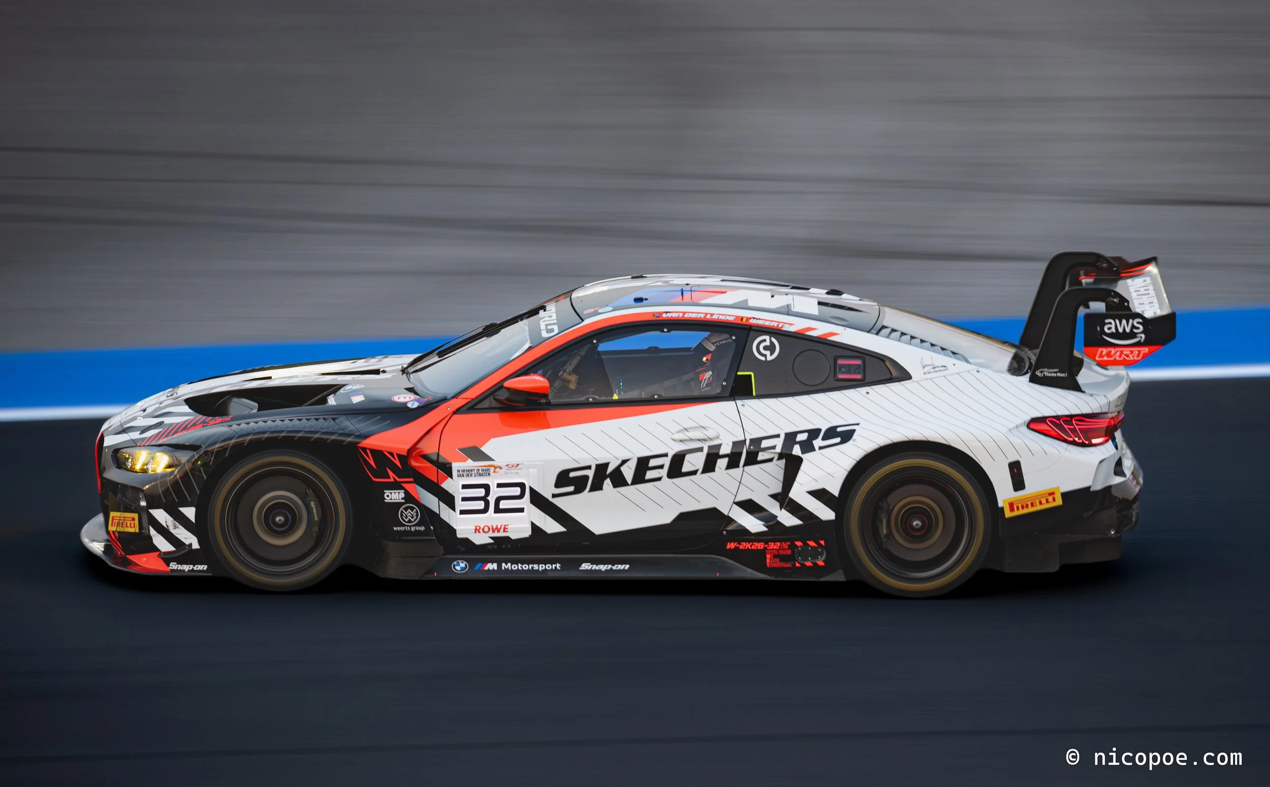 A race car with the number 32, pink, black, and white design, and various sponsor logos, driving on a race track with a grey wall in the background.