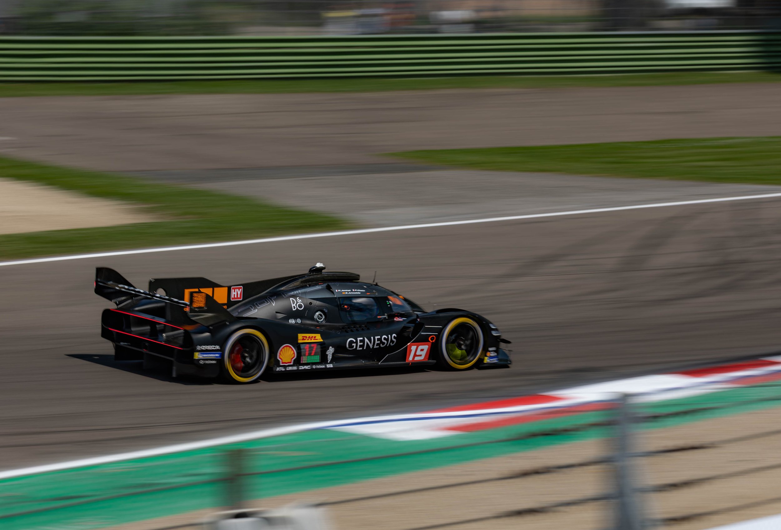 A sleek black race car with logos and sponsorship decals speeds along a race track, with a blurred background of green and grey barriers.