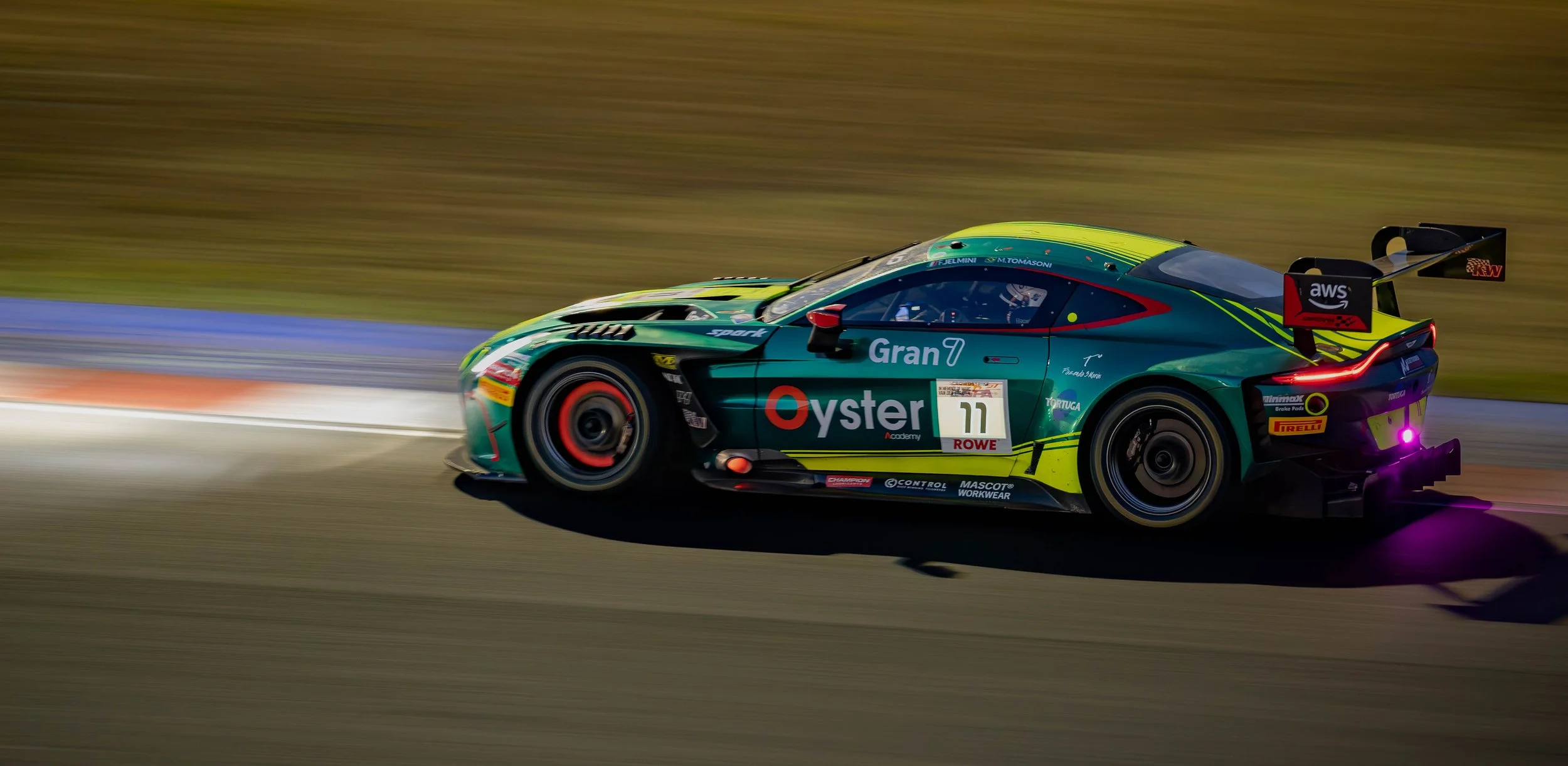 A green race car with various sponsor logos, including Oyster, Pirelli, and Rowe, speeding on a racetrack at night, producing motion blur.
