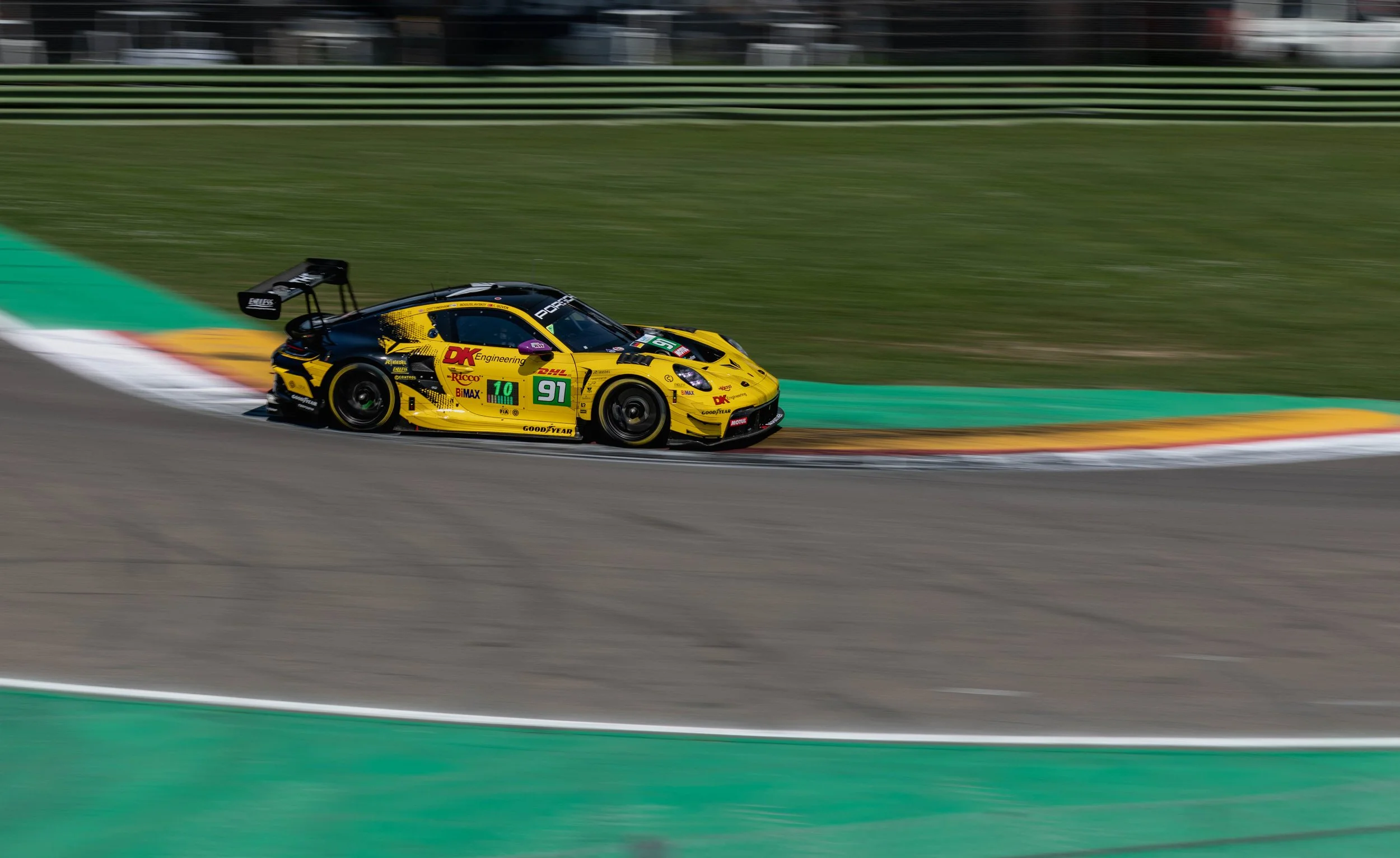 A yellow and black race car speeding on a race track with visible motion blur, surrounded by green and yellow curbing.