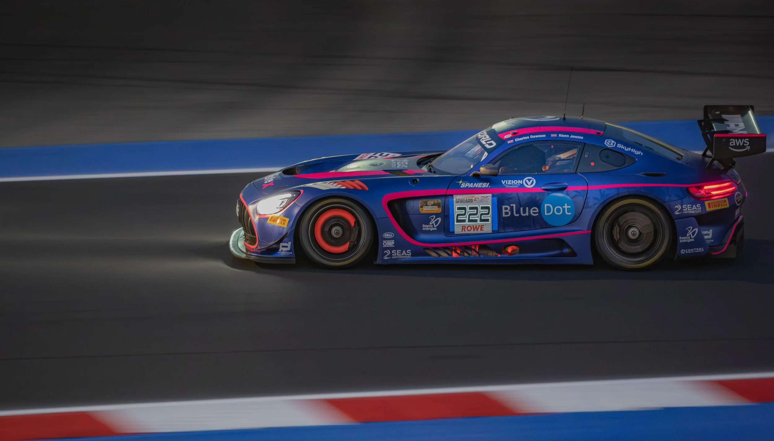 A blue race car with pink accents driving on a racetrack during nighttime, with sponsor logos and the number 222 on the side.