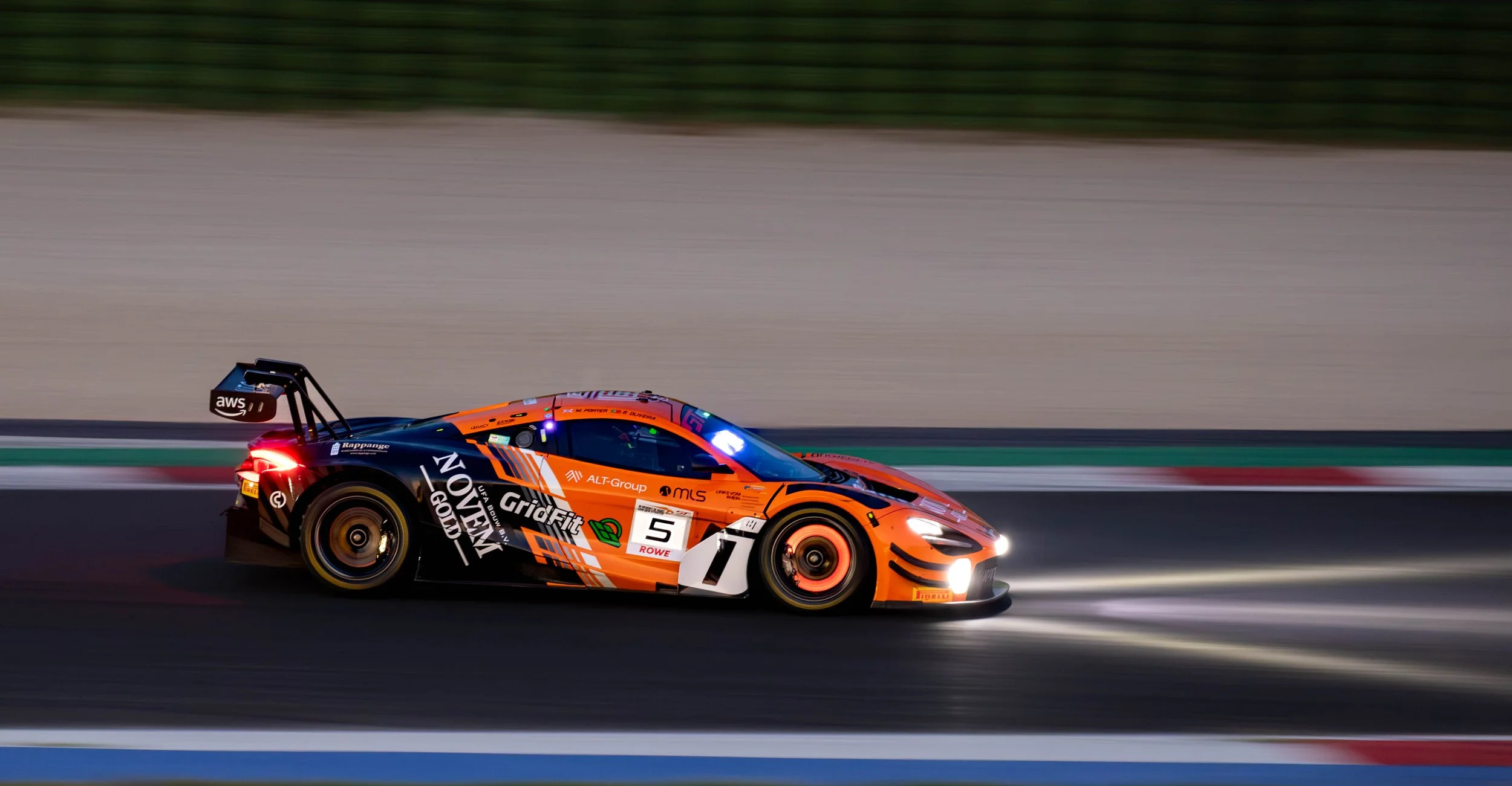 An orange and black racing car speeding on a racetrack with a blurred background, featuring sponsorship logos including 'GridFit' and 'AWS'.