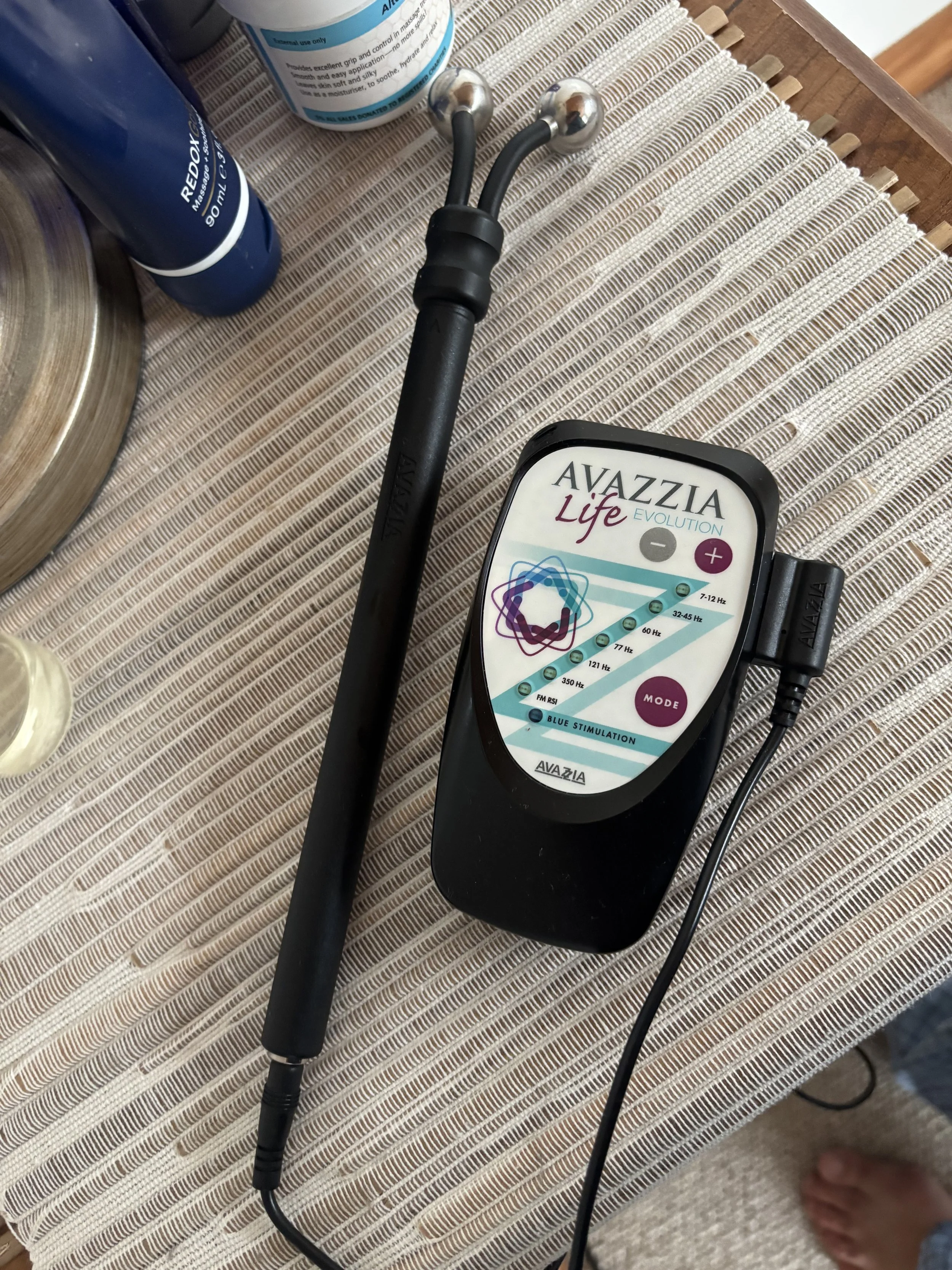A black handheld device with a colorful label reading 'Avazzia Life Evolution' connected to a black cable, placed on a woven beige tablecloth. The device has a dial and buttons, and a metal fumace connected to it.