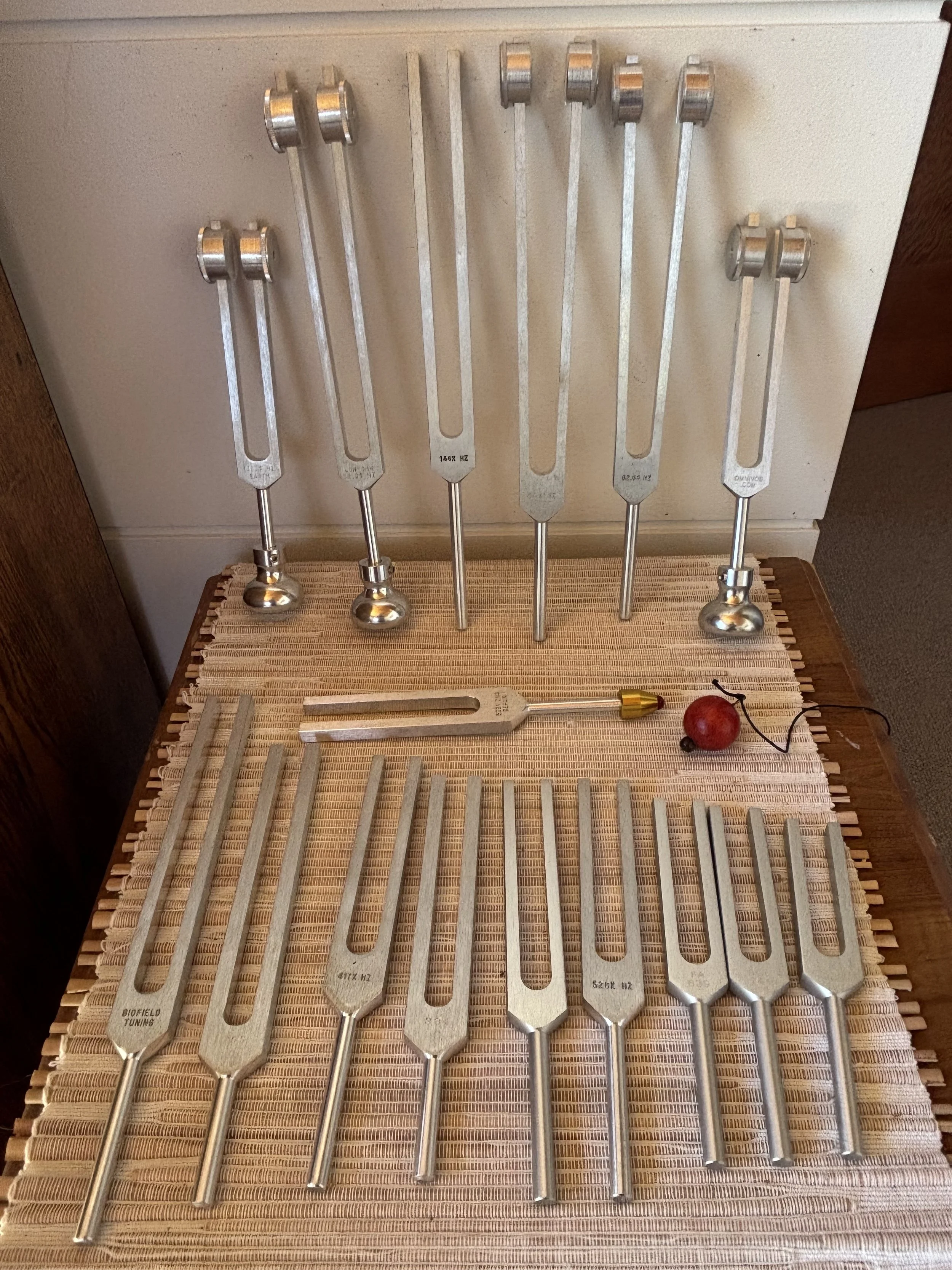 Collection of tuning forks of various sizes and frequencies, arranged on a woven mat on a wooden surface, with a small wooden handle, a red artificial berry, and a thin wire with a paper clip attached.