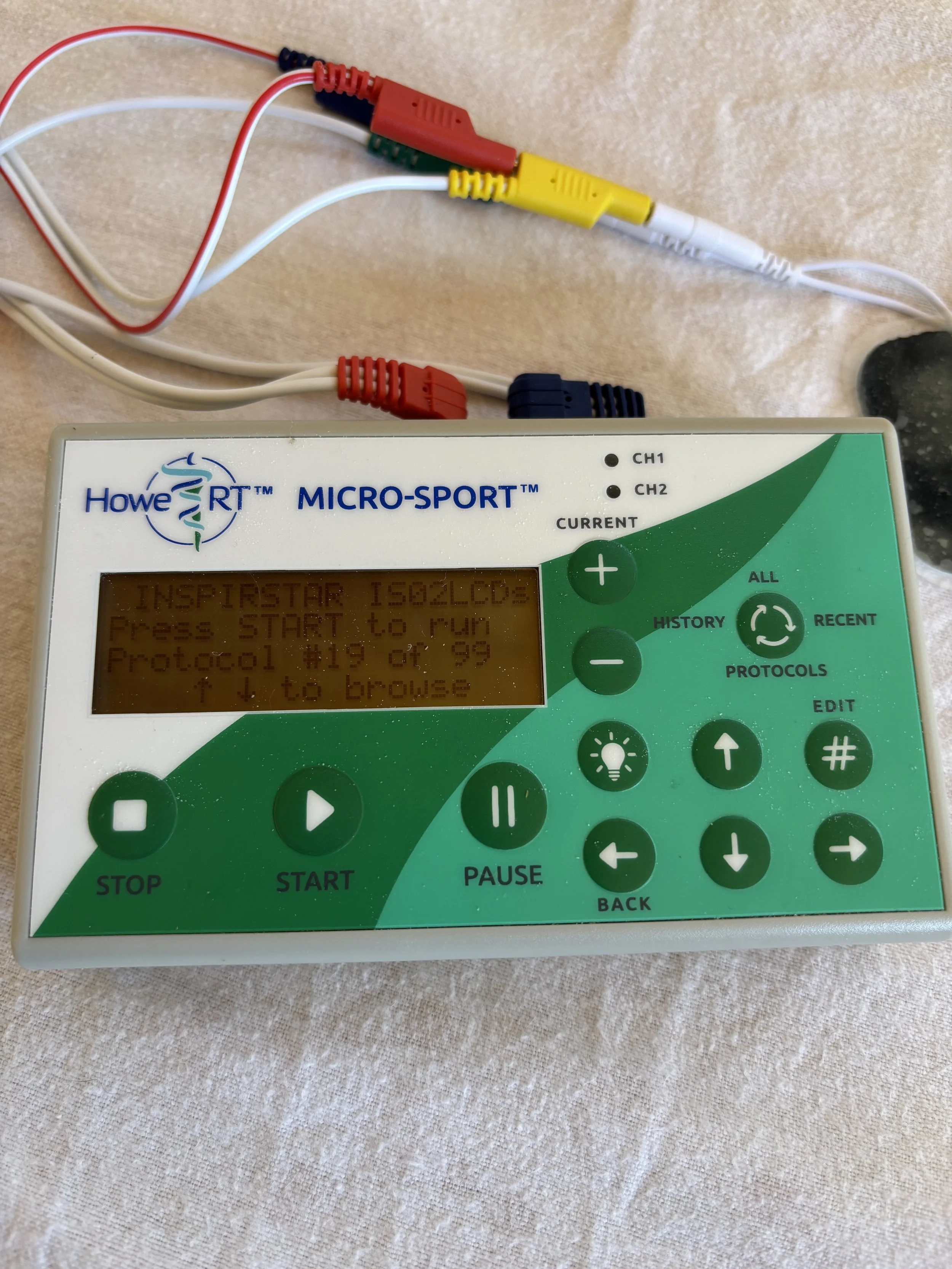 A microcontroller device called Howe RT MICRO-SPORT with wires connected to its top, situated on a beige surface. The device has an orange, backlit LCD screen displaying text and a green control panel with buttons for various functions.