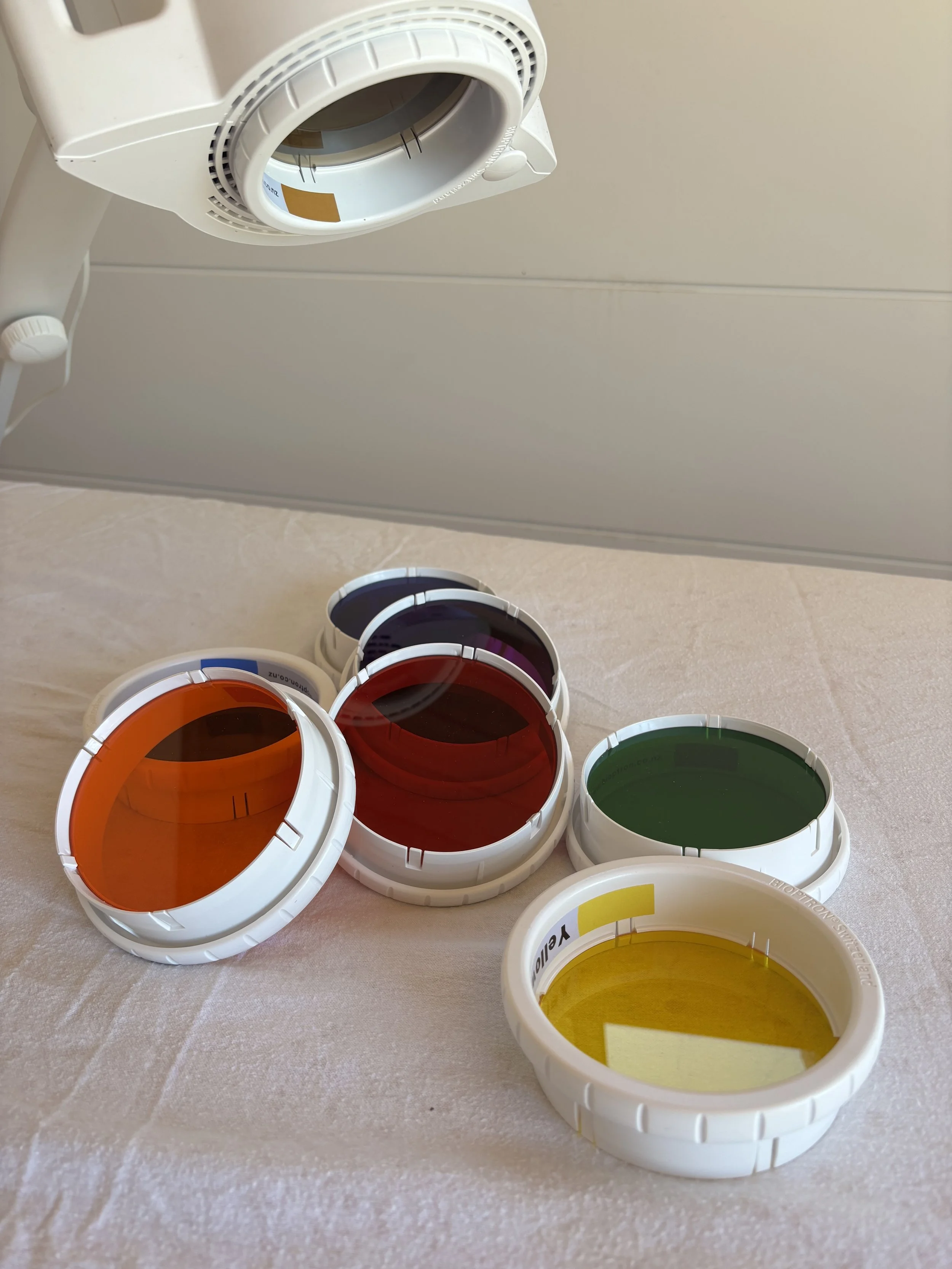 A collection of colored contact lenses in various colors including orange, red, purple, green, and yellow, displayed on a light-colored surface with a sheet covering it. In the background, an indoor ceiling with a white vent and part of a ceiling-mounted device is visible.
