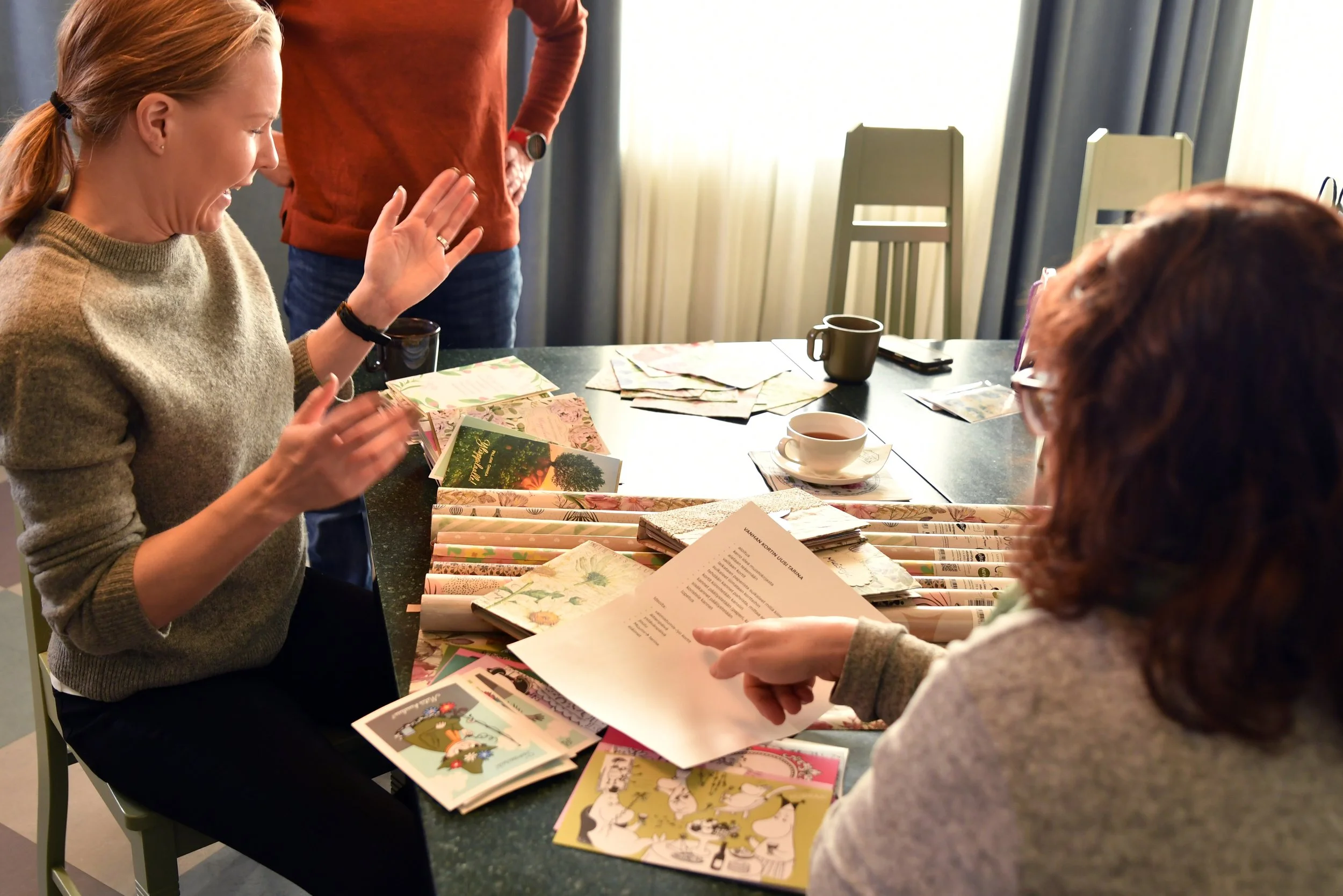 Suvi Nisula yhdessä kahden Suomen Korttipisteen työntekijän kanssa keskustelevat ja katsovat pöydällä olevia värikkäitä kortteja ja papereita, joita he ovat kuvaamassa askarteluvideota varten.