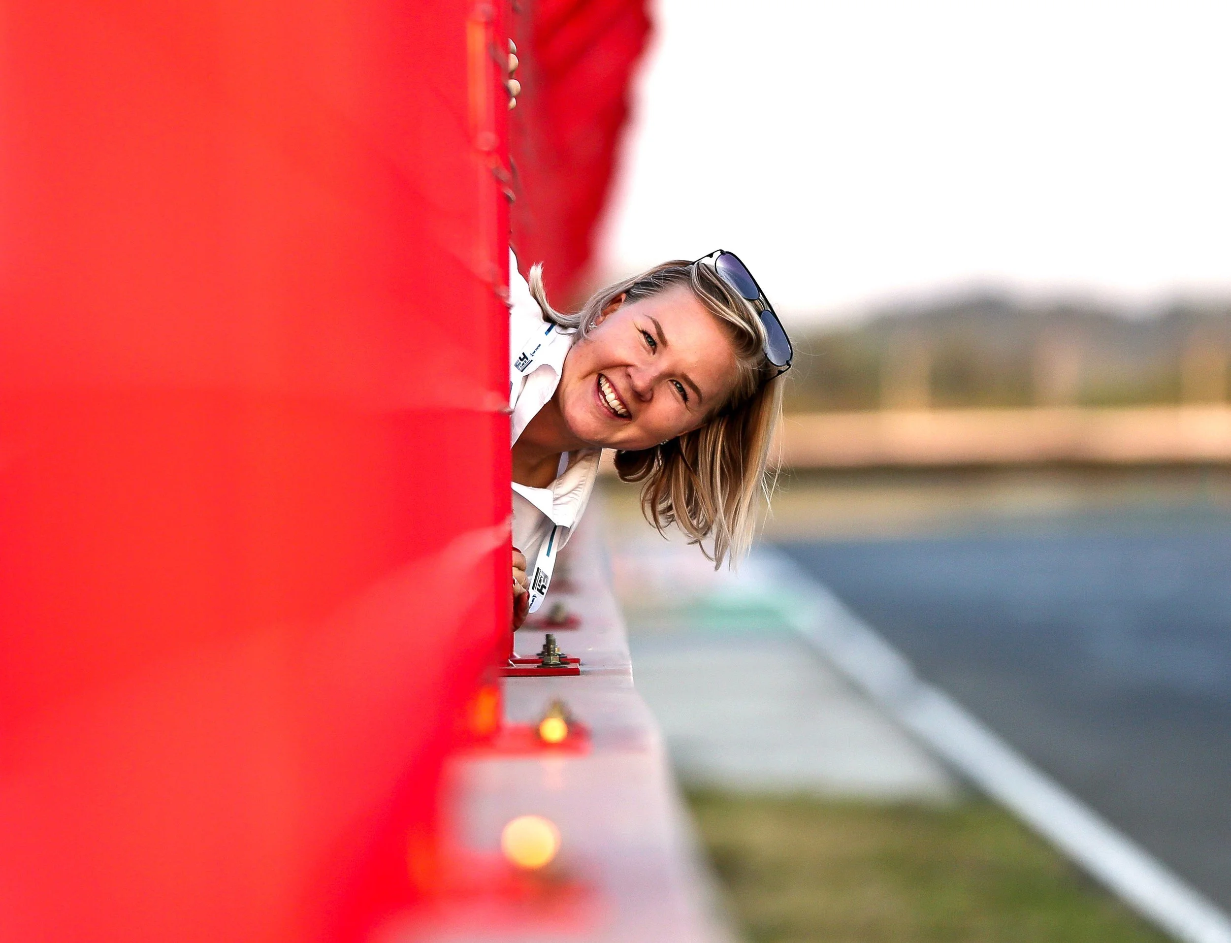 Suvi Nisula kurkkaa moottoriradan pitwallin verkon välistä ja hymyilee kohti kameraa, ilta-auringon sarastaessa.
