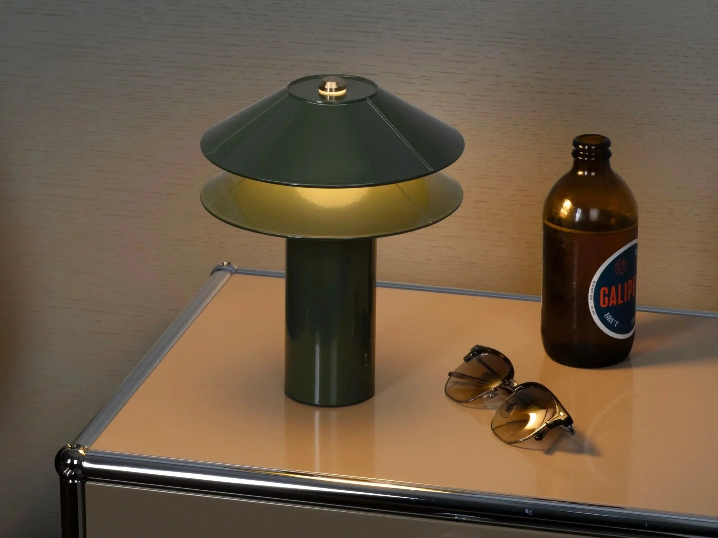 A table lamp with olive green shades, a brown glass bottle with a blue and red label reading 'Galip', and a pair of sunglasses with yellow-tinted lenses resting on a beige table.
