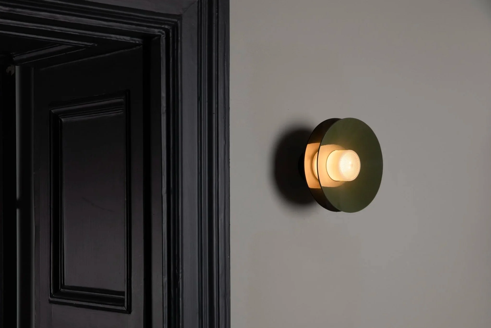 A modern wall sconce light with a circular, dark green or black shade and a soft glowing bulb, mounted on a light-colored wall near a black paneled door.