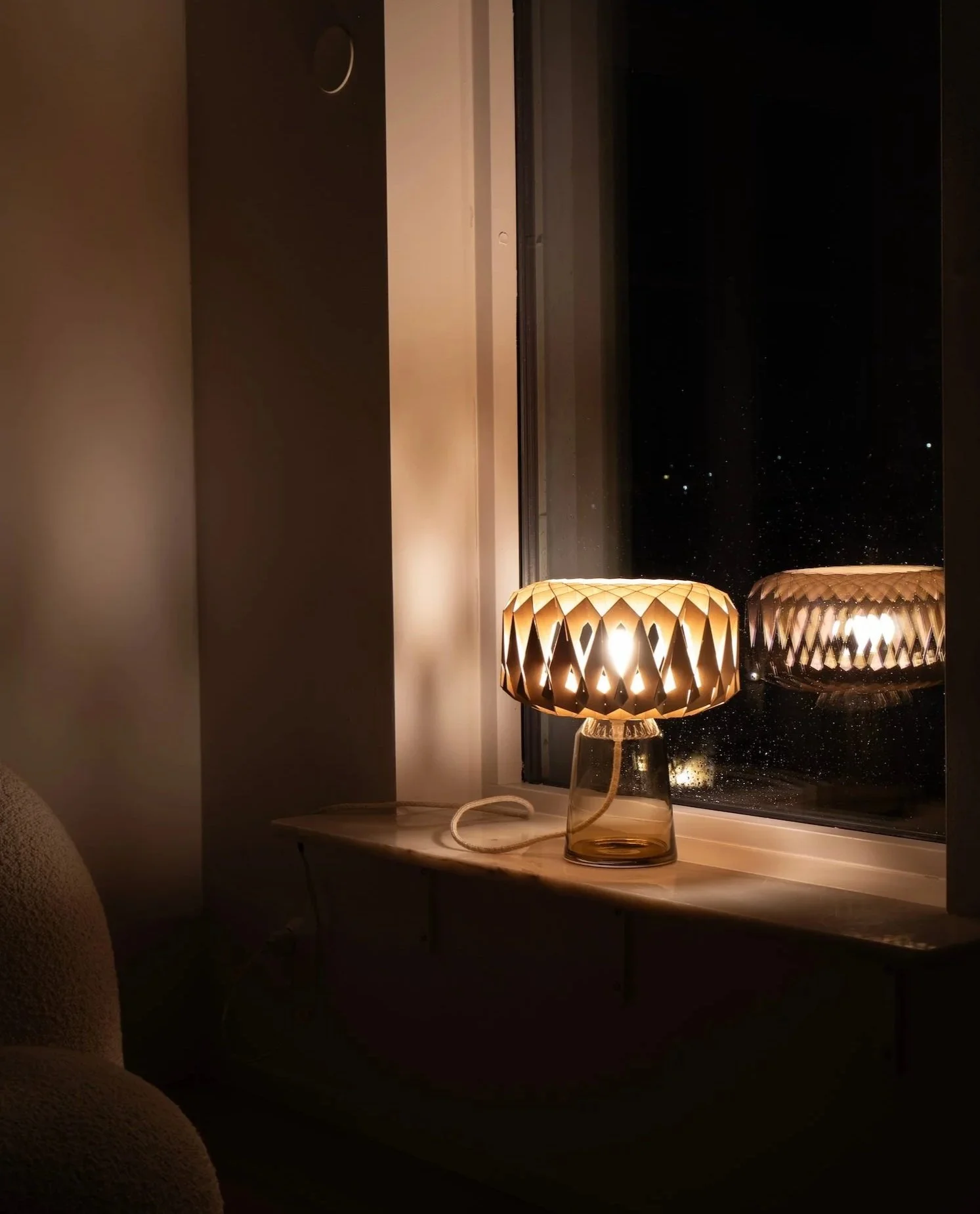 A lit decorative lamp on a windowsill at night, with the lamp's reflection visible on the window glass.