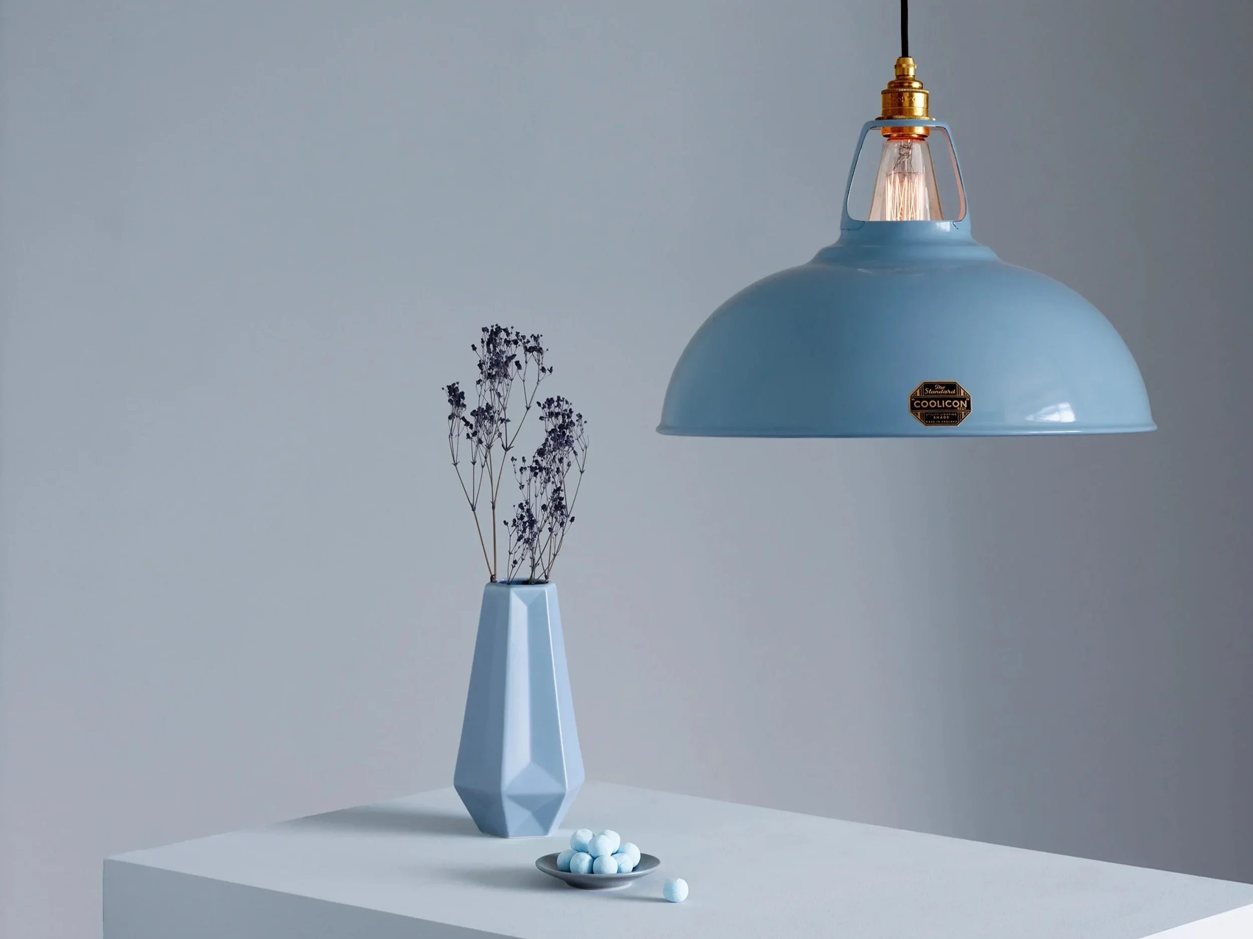A blue pendant lamp hanging over a white table with a pale blue geometric vase holding dried purple flowers and a small plate of white and light blue candies in front.