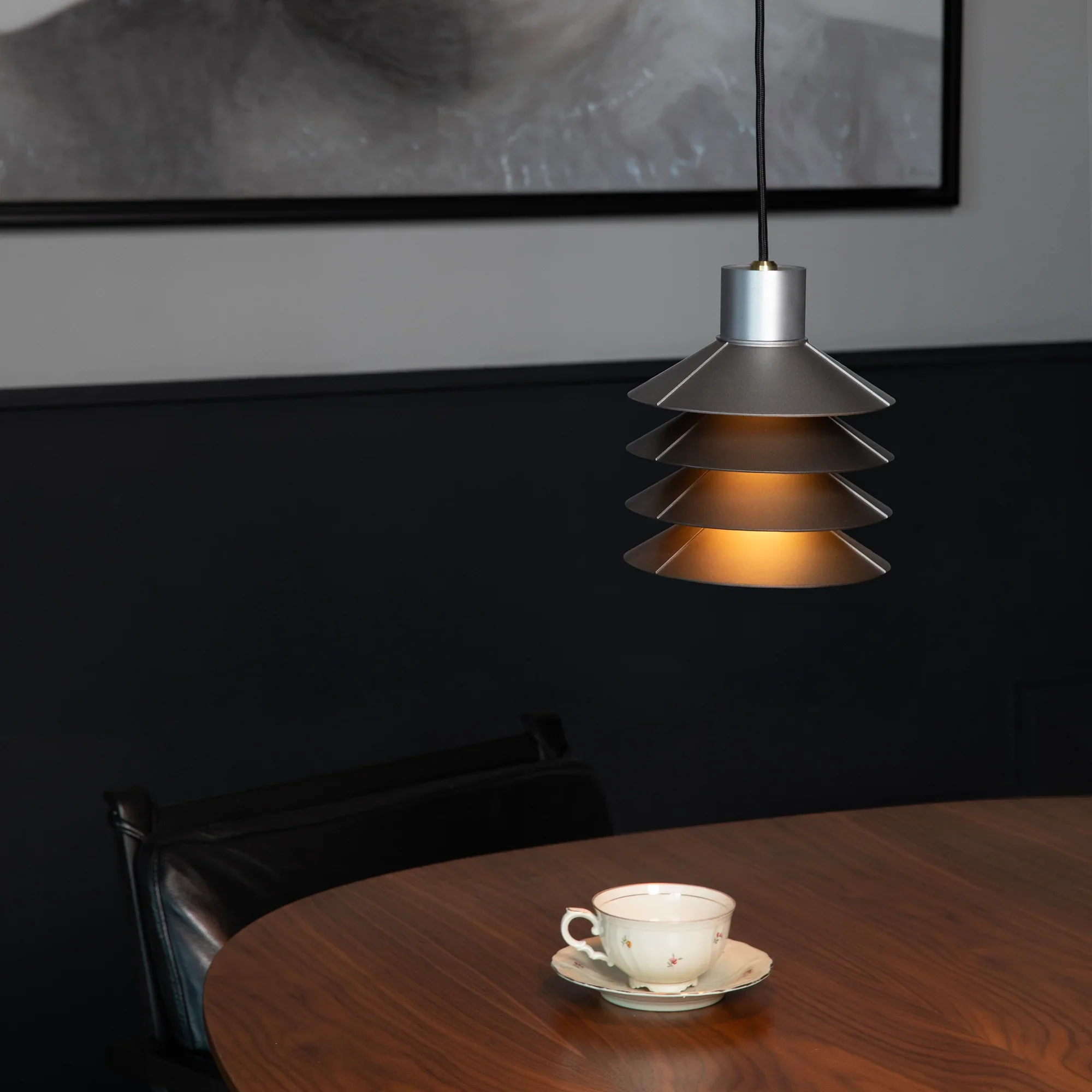 A modern pendant lamp with a layered, metallic design hanging over a wooden dining table with a china teacup and saucer.